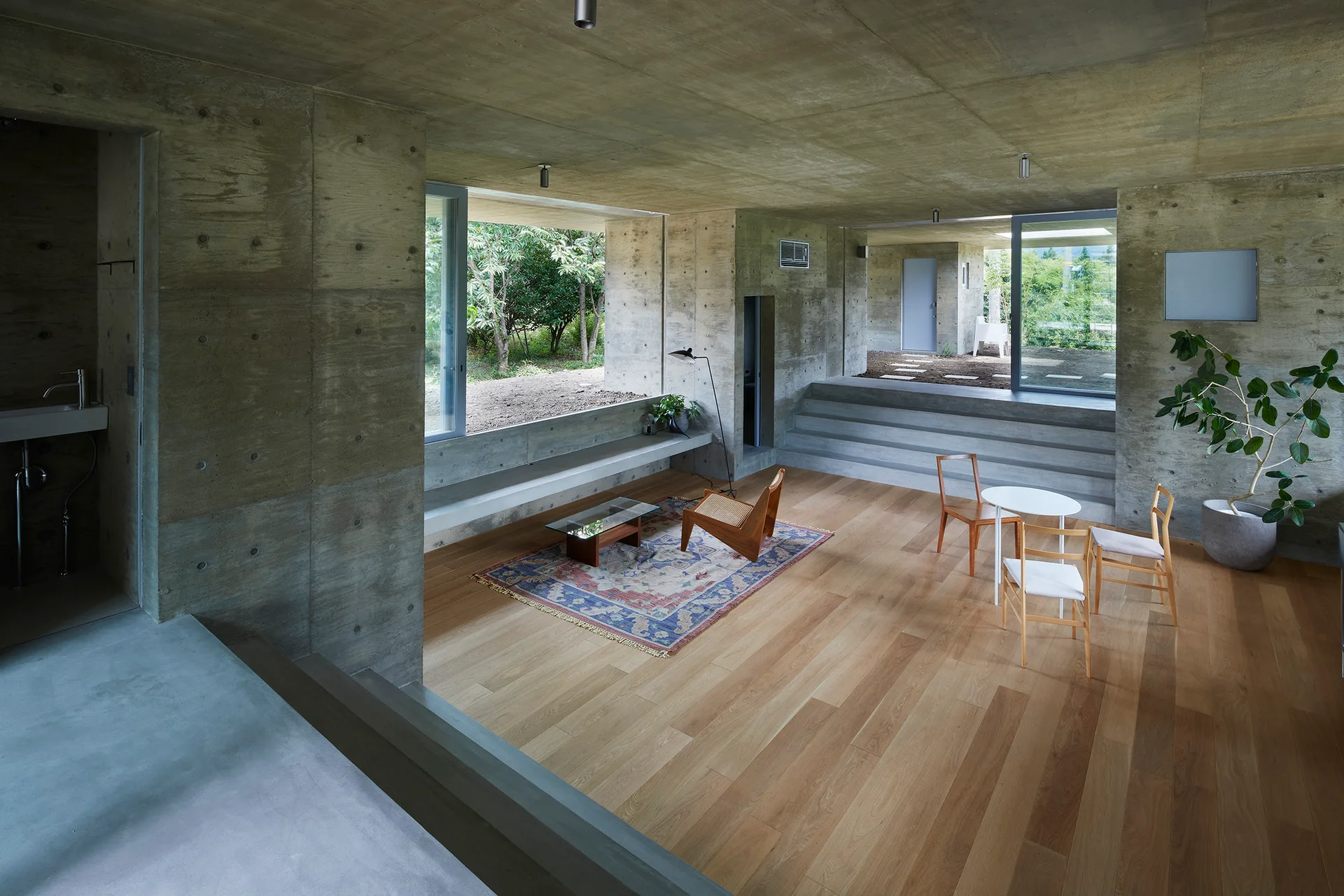 A minimalist living room features raw board-formed concrete walls, natural wood plank flooring, a small dining set, and an ornate area rug.