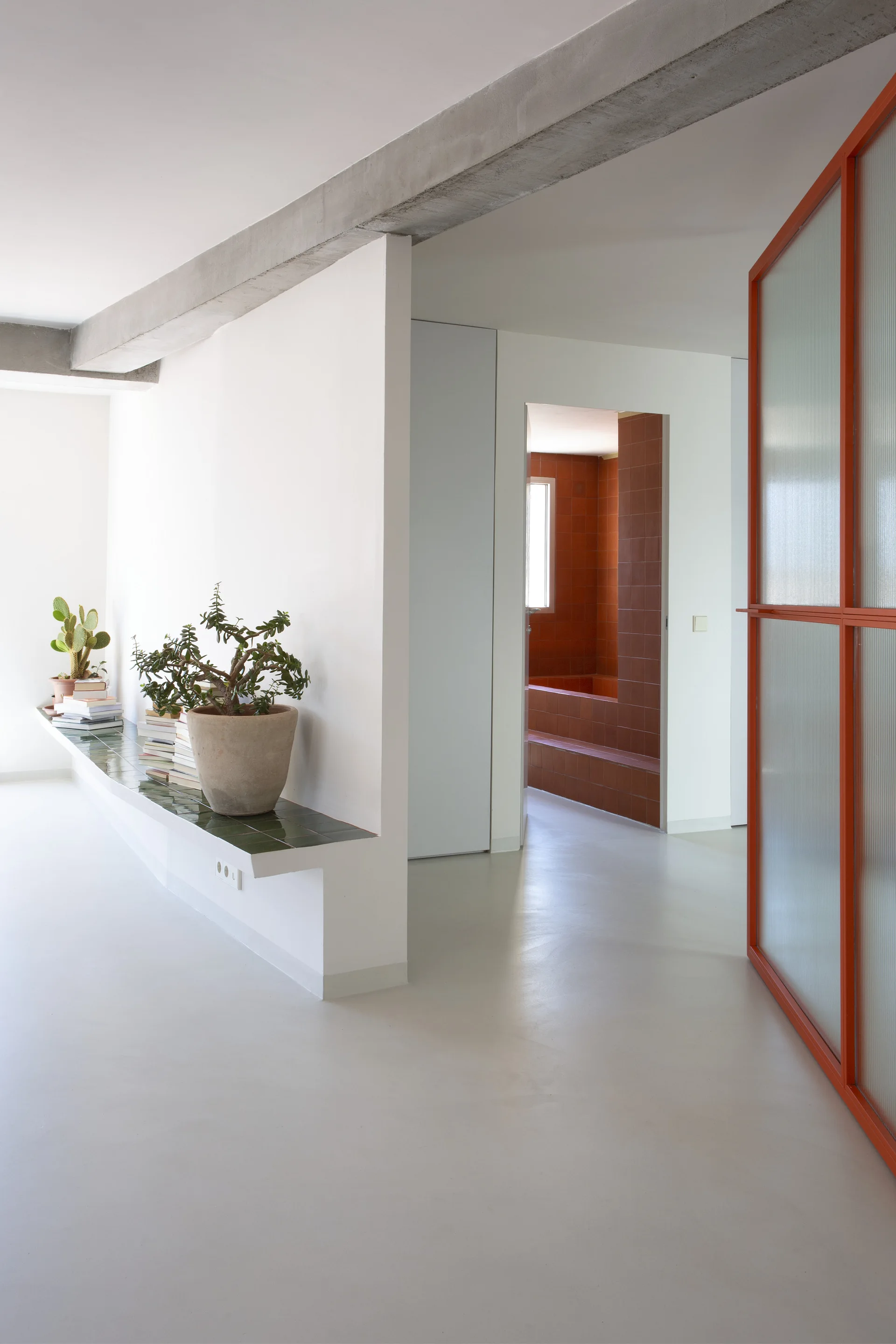 A minimalist white hallway features a low built-in bench topped with green ceramic tiles, an exposed concrete beam, and a red-framed glass sliding door.