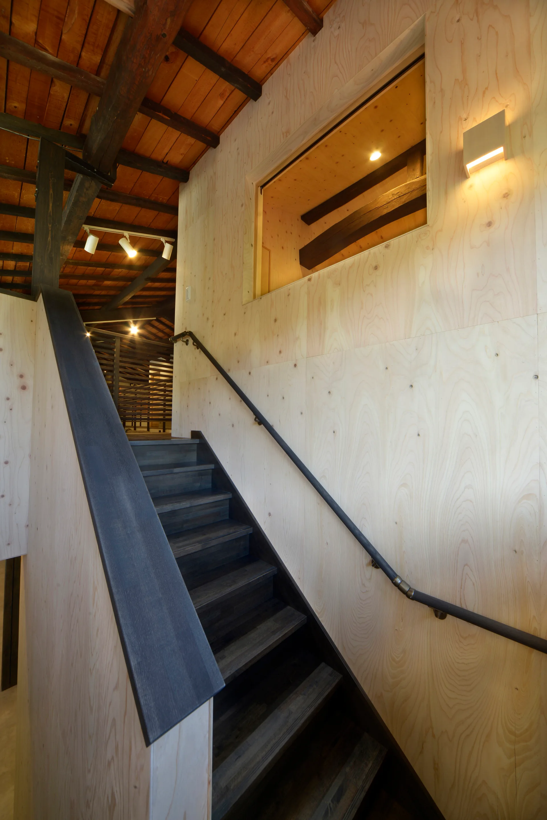 A wooden staircase with dark stained treads rises against light plywood-paneled walls under an exposed dark wood beam ceiling.