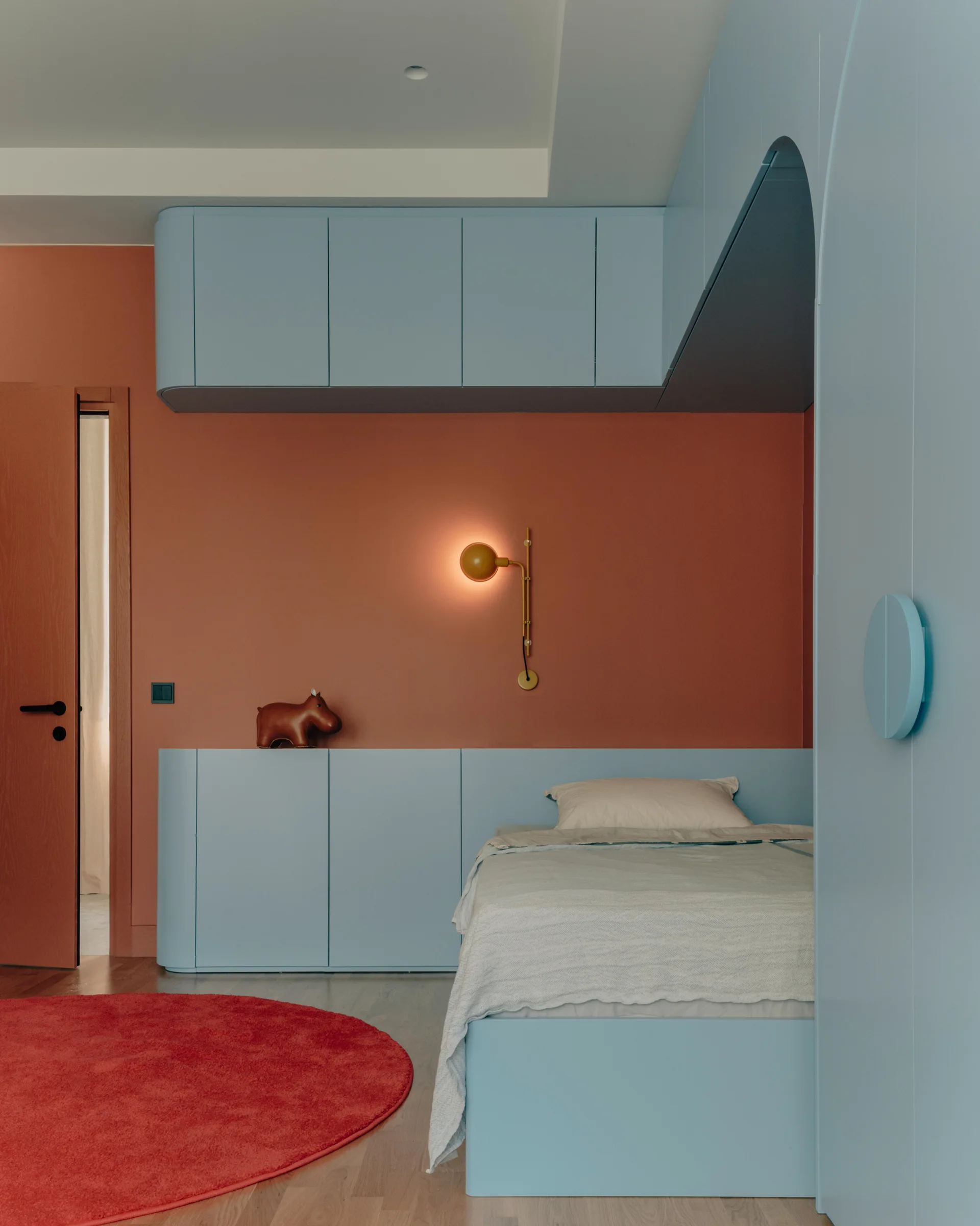 A modern bedroom features terracotta walls, light blue custom built-in cabinets framing a bed, a round red rug, and a glowing globe wall sconce.
