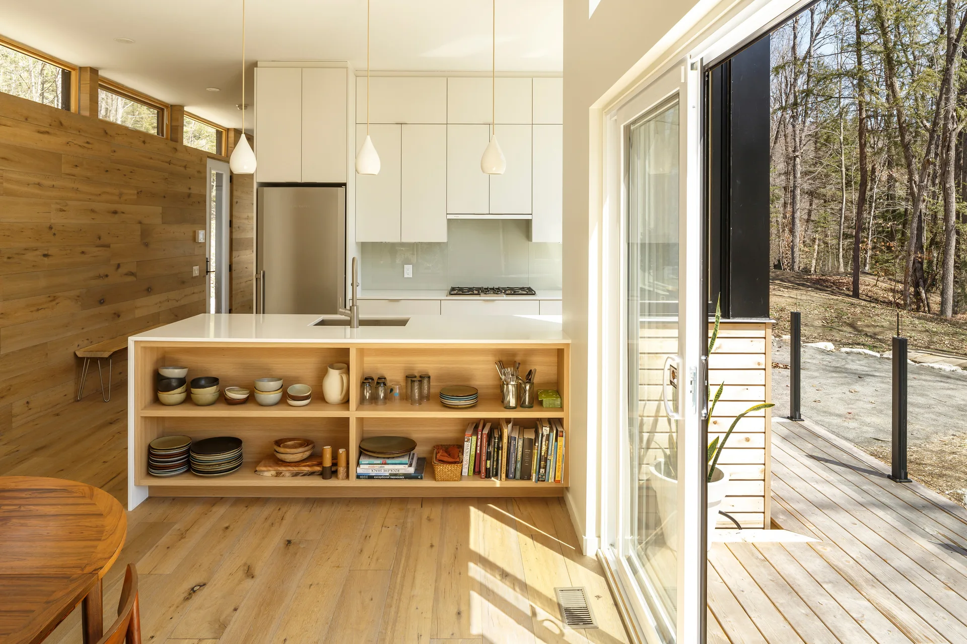 A modern kitchen features white cabinetry, a light wood island with open shelving, and a sliding glass door leading to an outdoor deck.