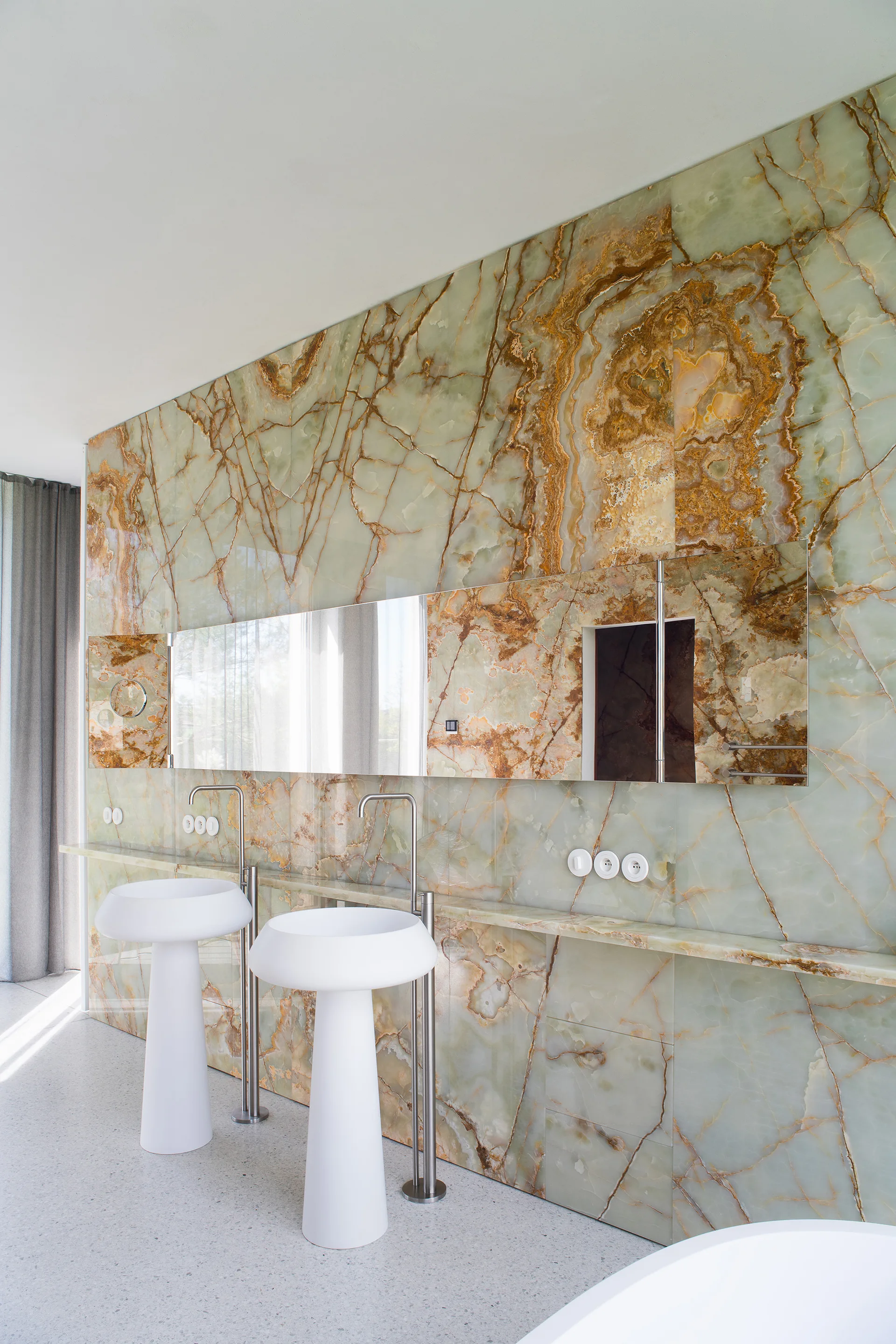 Two white pedestal sinks stand before a highly veined green onyx accent wall with an integrated horizontal mirror.