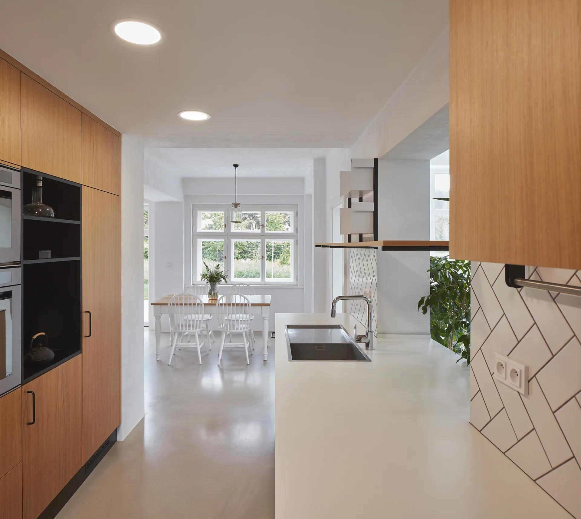 A modern kitchen features wood cabinetry, a white countertop with a sink, a herringbone tile backsplash, and a dining table with white chairs.