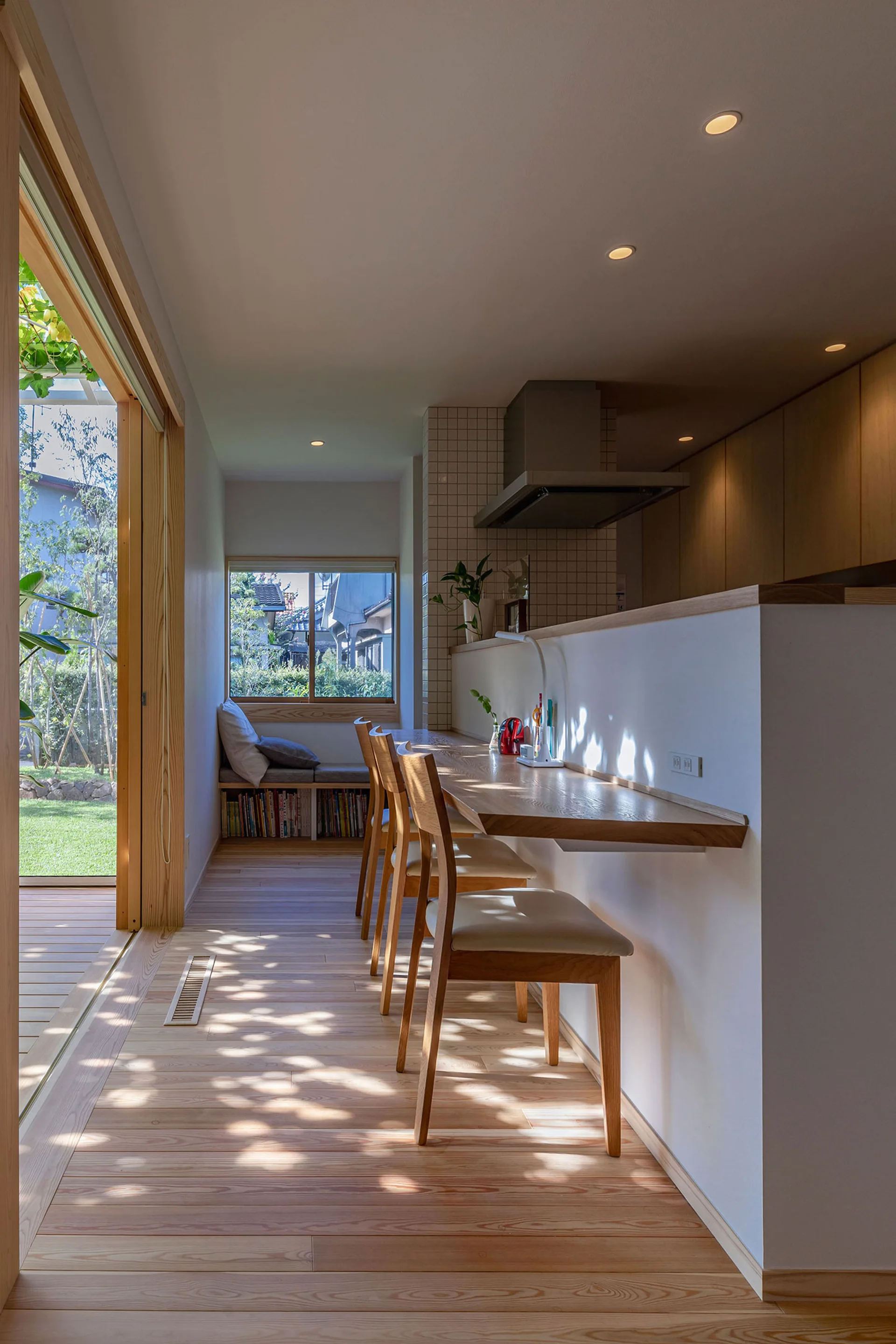 A modern light wood dining area featuring a floating bar counter with chairs, a window seat, and sliding glass doors opening to a green garden.