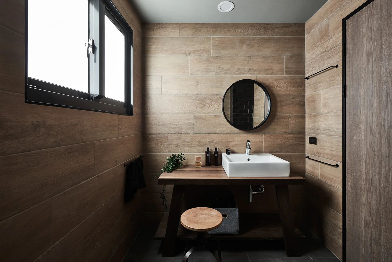A modern bathroom features horizontal wood-look wall tiles, a dark wood table vanity with a white vessel sink, and a round black-framed mirror.