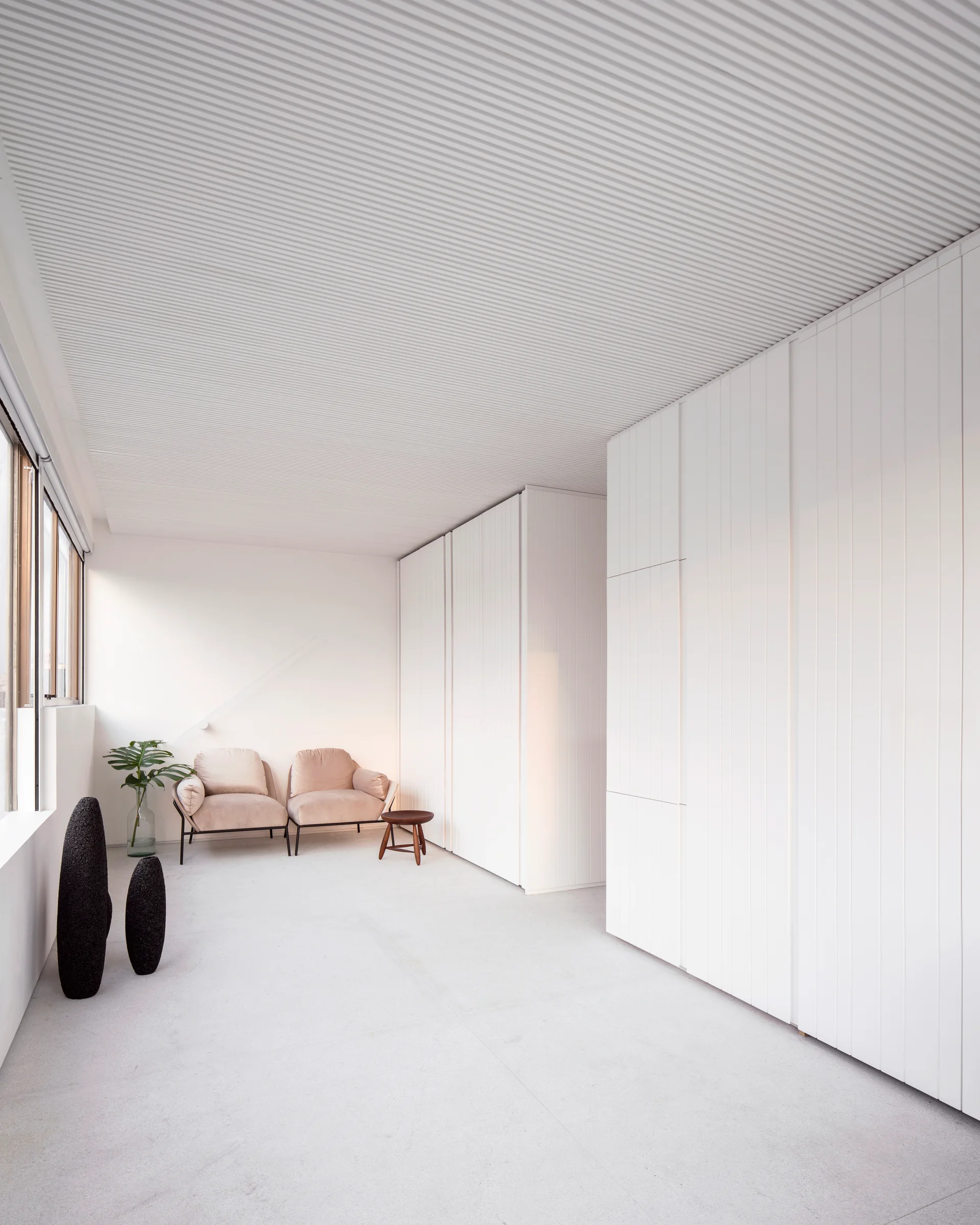 A minimalist white bedroom featuring a ribbed ceiling, floor-to-ceiling cabinetry, two pale pink armchairs, and two tall dark sculptures.