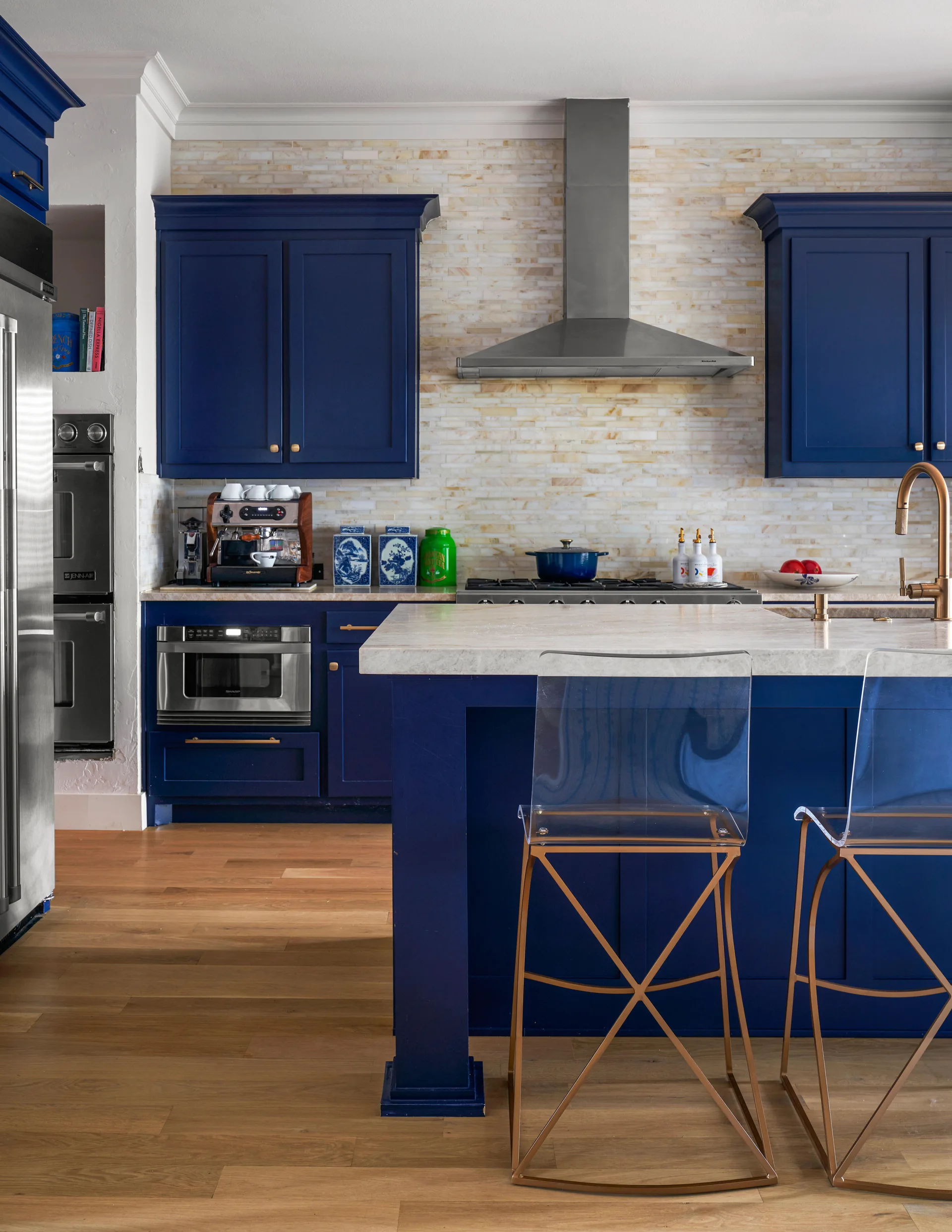 A modern classic kitchen features navy blue cabinetry, a stone-topped island with two clear acrylic bar stools, and a textured stone tile backsplash.