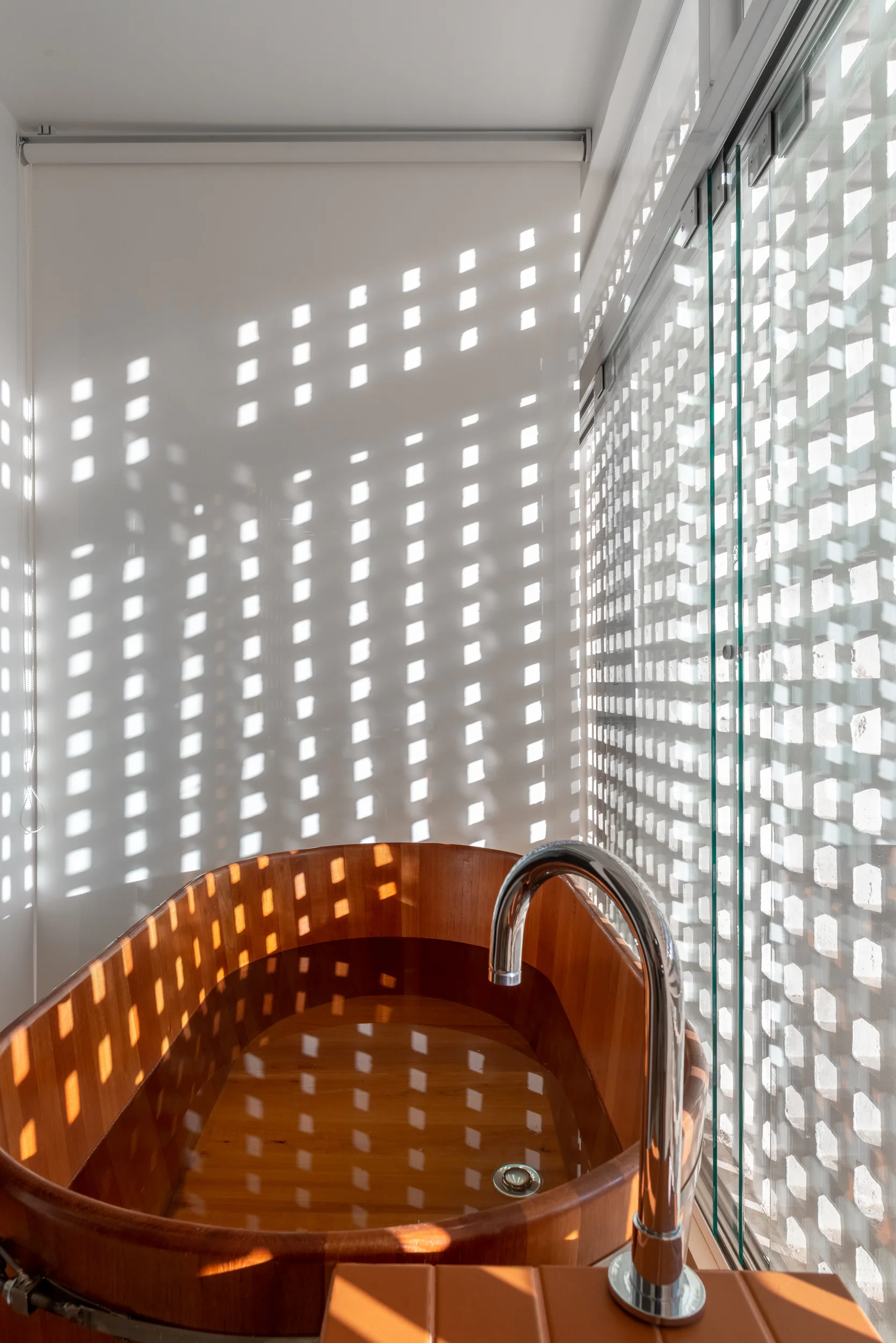 A wooden soaking tub is filled with water next to a glass window with a grid screen casting square shadows on a white wall.
