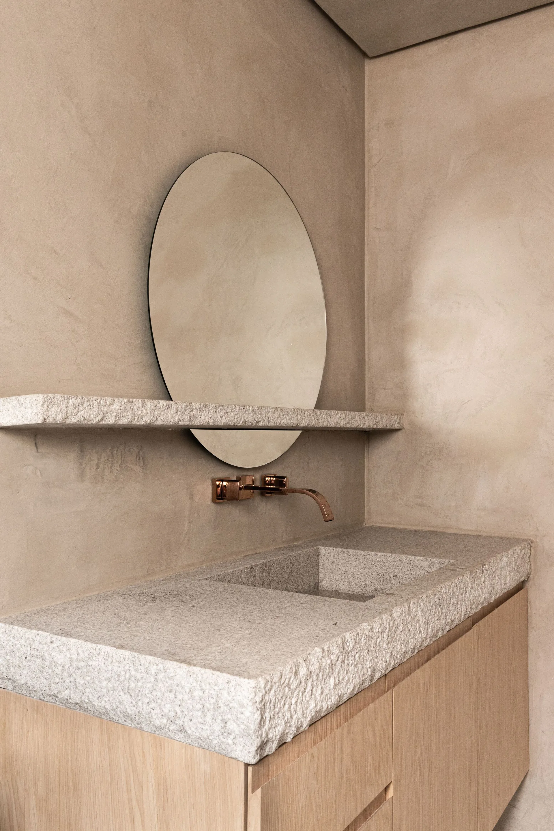A circular mirror is bisected by a floating stone shelf above a vanity with a rough-cut stone integrated sink and copper faucet.