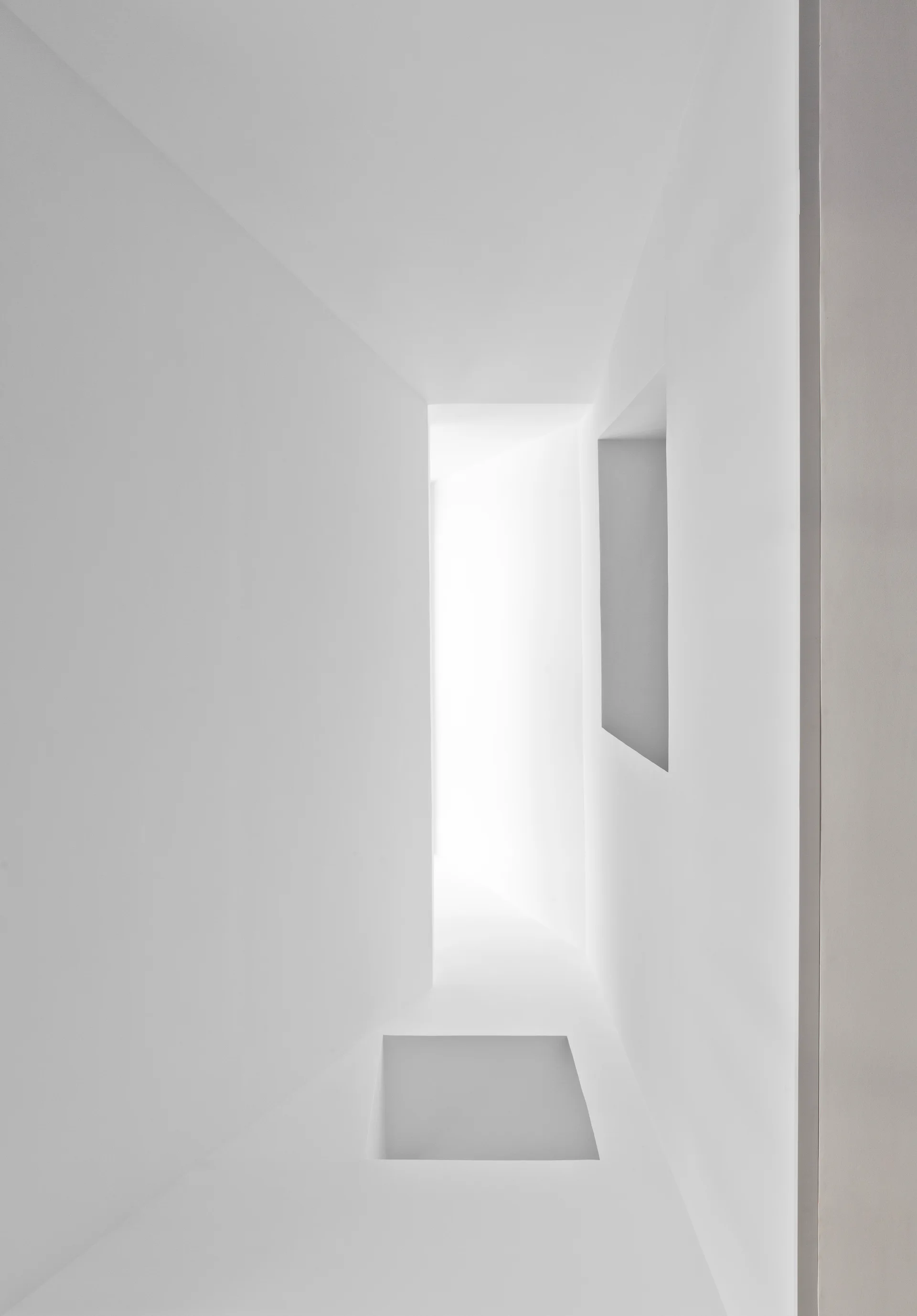 A minimalist white corridor features a recessed wall niche, sharp geometric angles, and bright natural light shining from the far end.