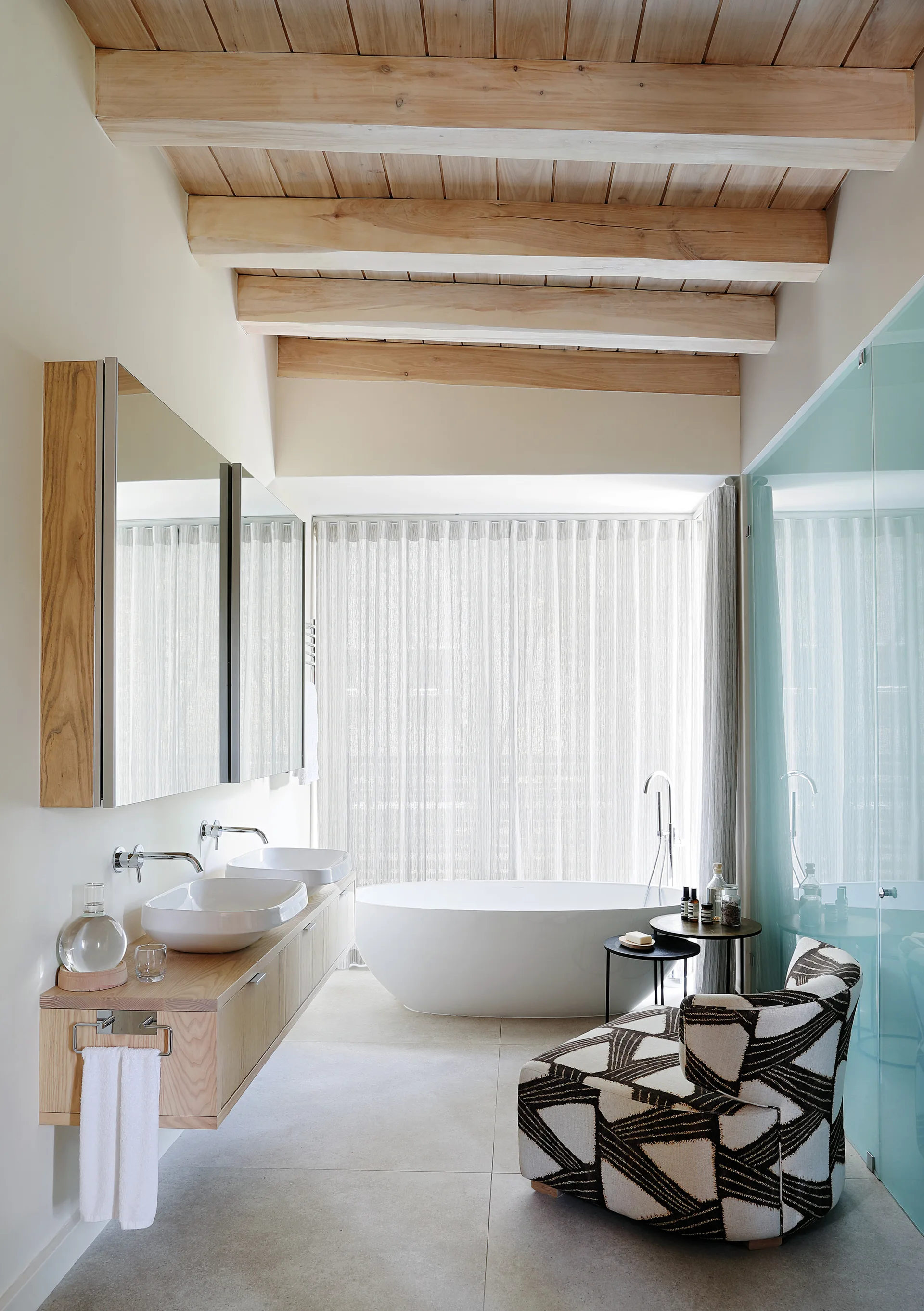 A modern bathroom features a floating wooden vanity with double vessel sinks, a white freestanding tub near a sheer curtain, and exposed ceiling beams.