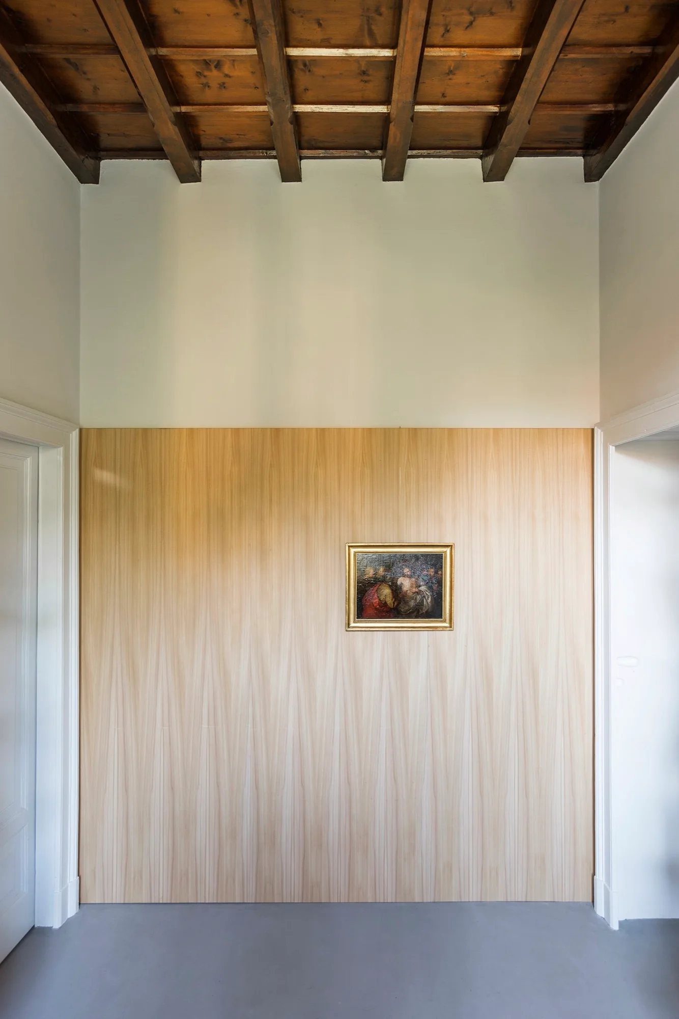 A modern hallway features a light wood paneled wall with a small framed painting beneath a dark wood beamed ceiling.