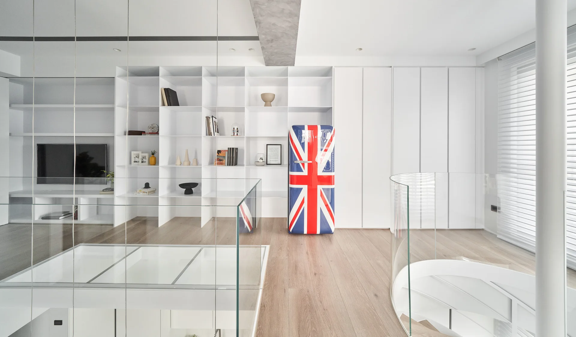 A bright landing featuring a Union Jack refrigerator alongside white built-in shelving, light wood floors, and a curved glass stair railing.