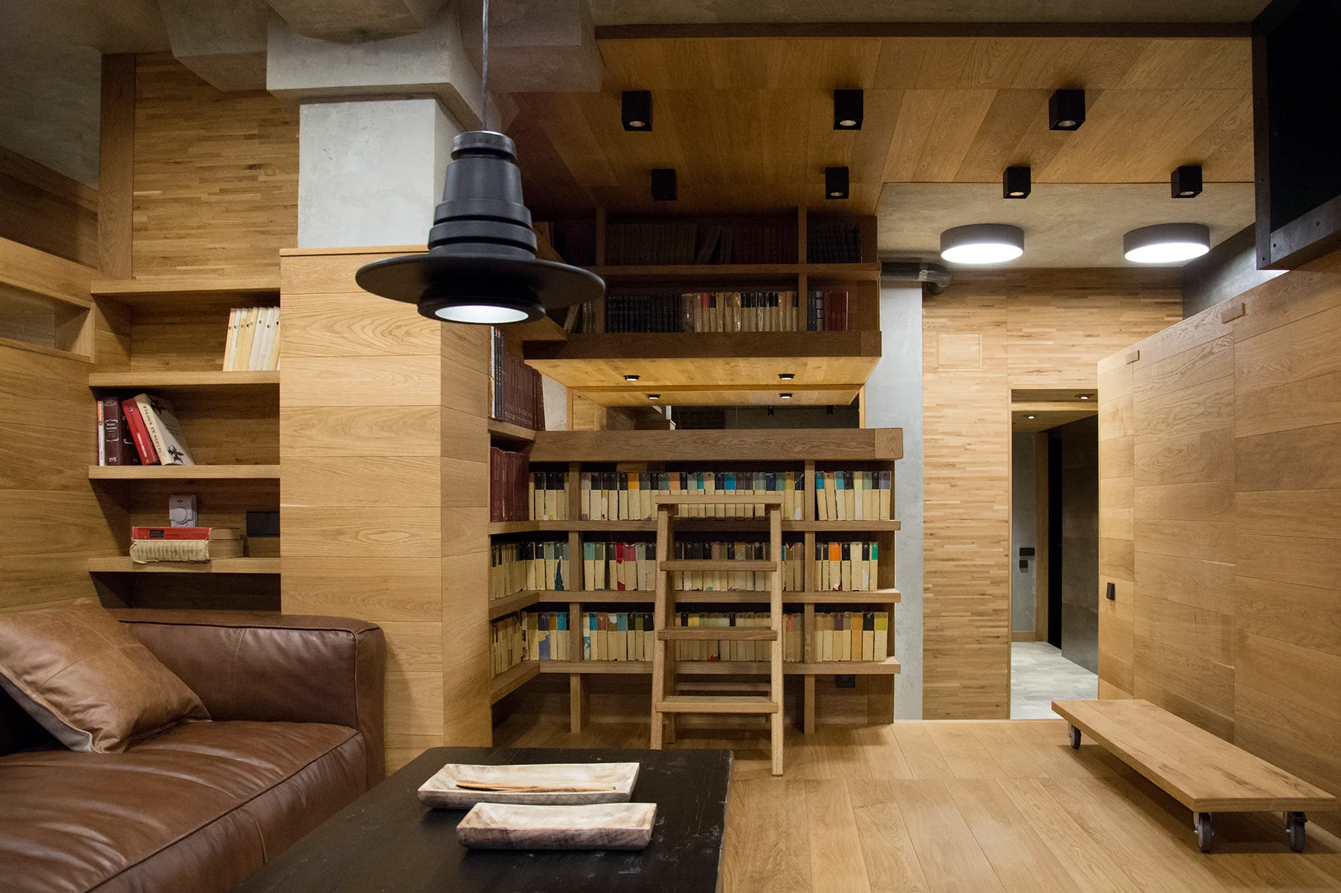 A home library featuring natural wood paneled walls and ceiling, matching built-in bookshelves with a ladder, and a brown leather sofa.