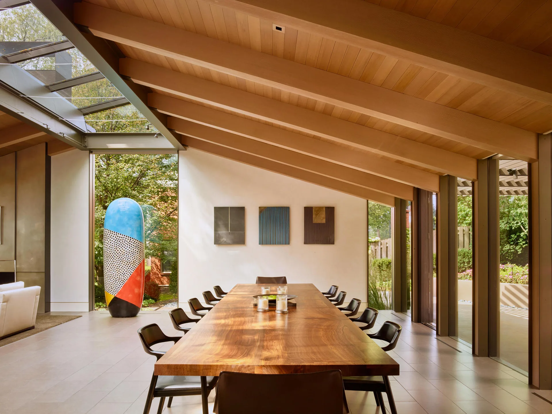 A spacious dining room features a long wooden table with black chairs beneath a sloped timber ceiling with exposed beams and glass walls.