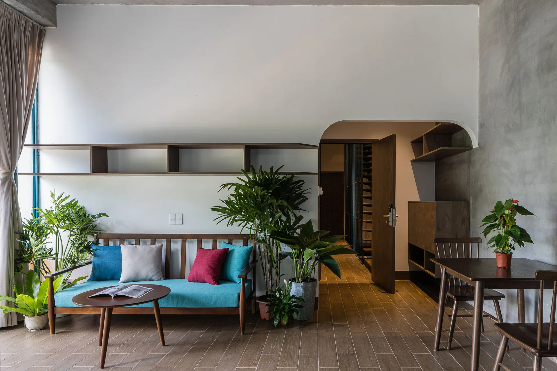 A modern living room features a teal cushion sofa, a round wooden coffee table, green potted plants, and an asymmetrical arched doorway.