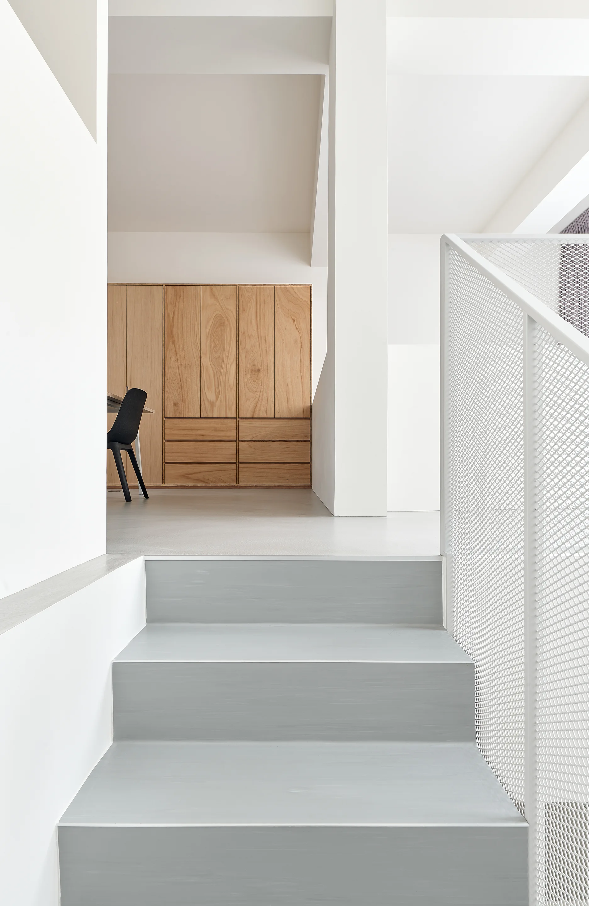 A minimalist gray staircase features a white metal mesh railing, leading to a landing with built-in light wood cabinetry and a black chair.