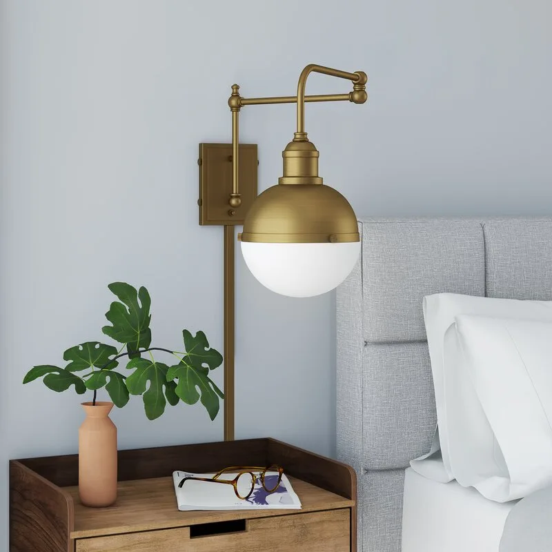 A brass swing-arm wall sconce with a white globe shade is mounted above a wood nightstand next to a gray upholstered headboard.