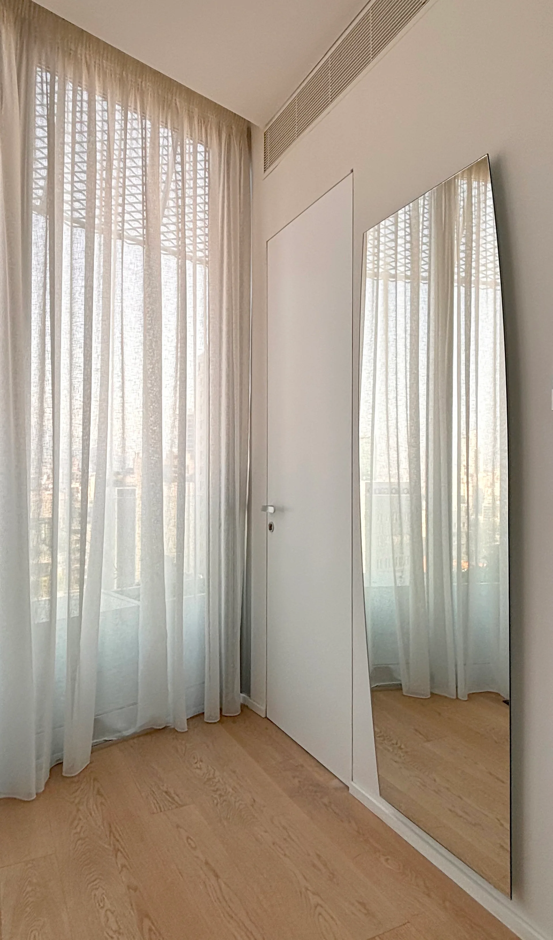 A minimalist interior space features floor-to-ceiling sheer white curtains, a flush white door, light wood floors, and a tall vertical wall mirror.