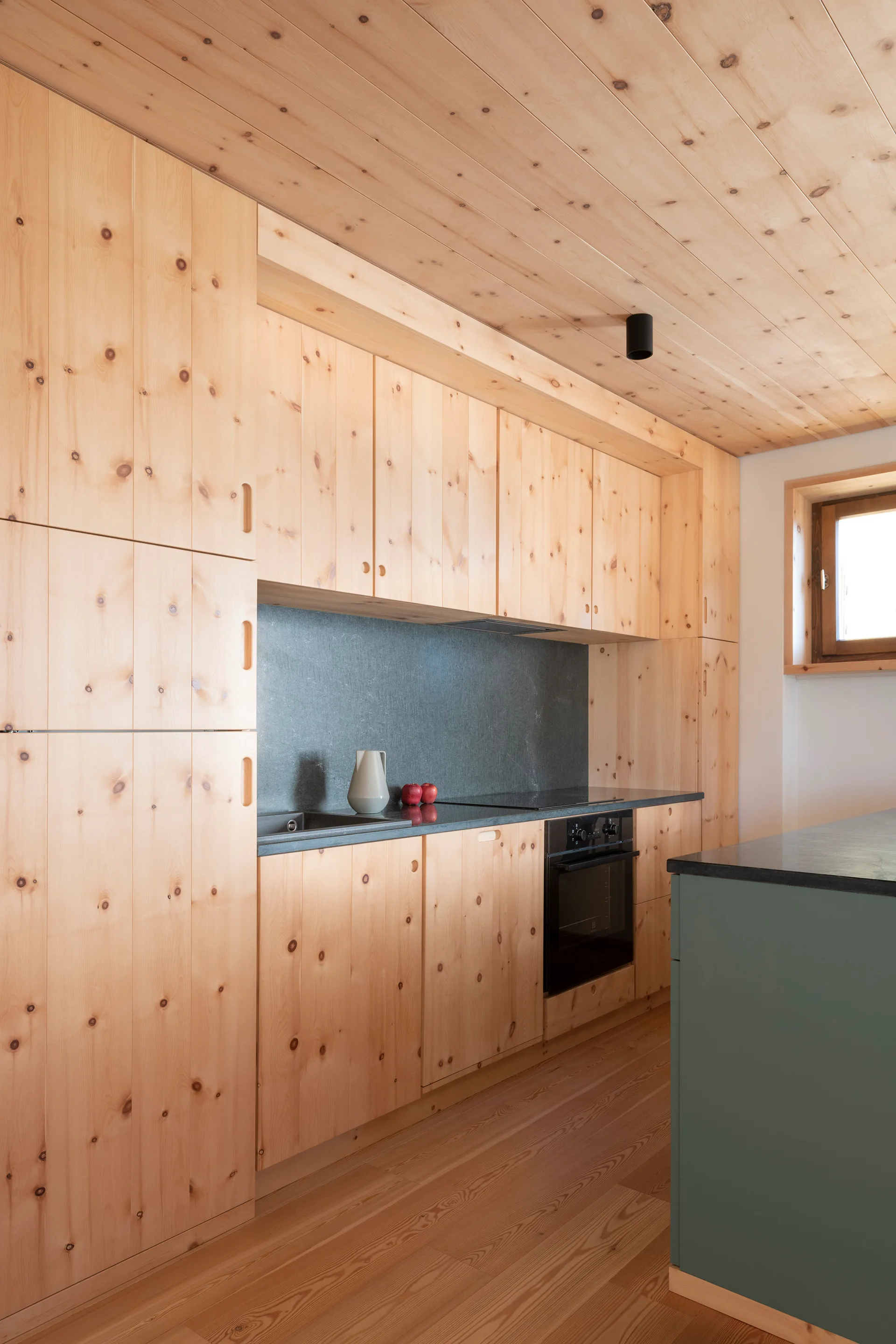 A minimalist kitchen featuring knotty pine wood paneled cabinets and ceiling, a dark stone backsplash, and a muted green island in the foreground.