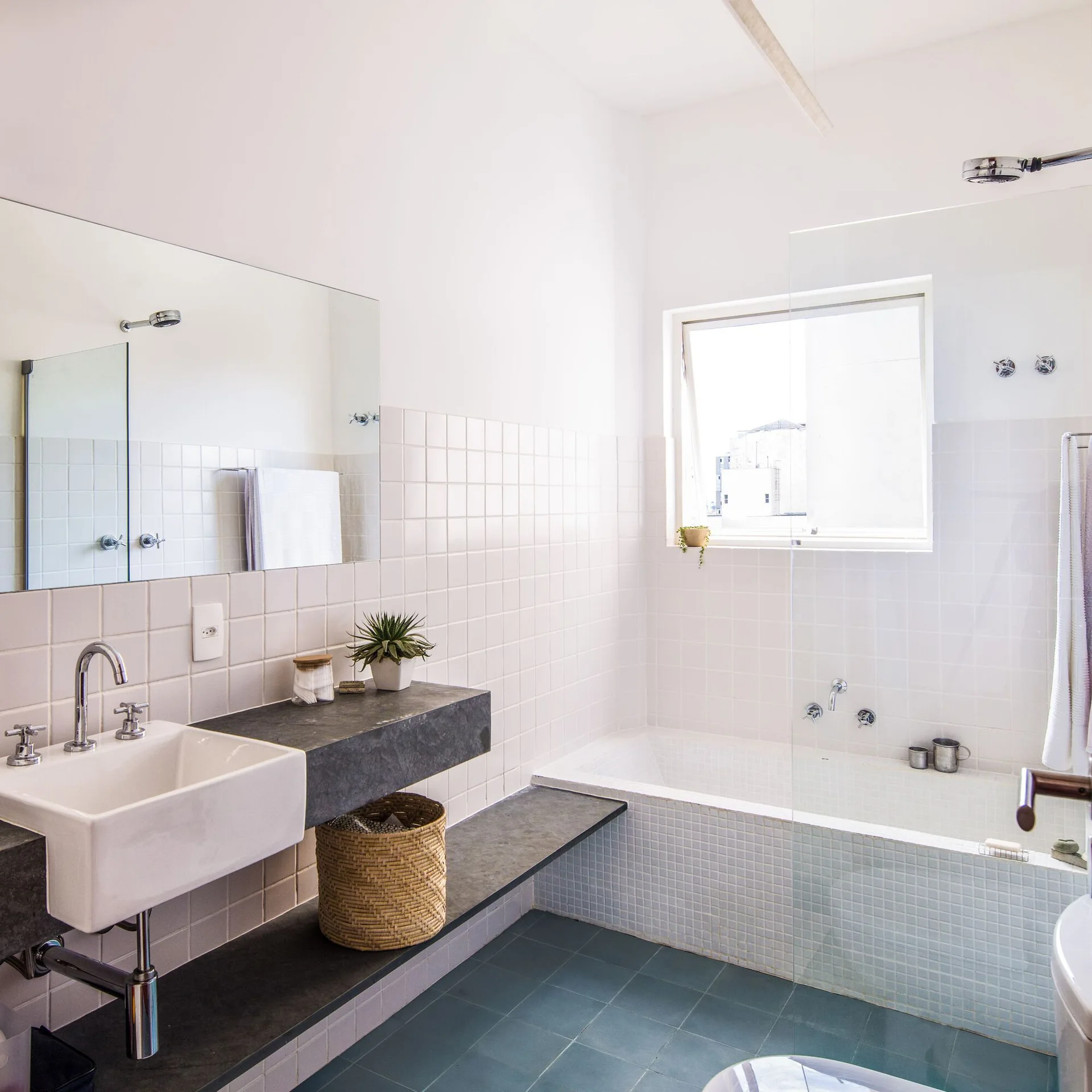 A minimalist bathroom features white grid-tiled walls, a wall-mounted sink on a dark stone counter, a tiled tub, and blue floor tiles.
