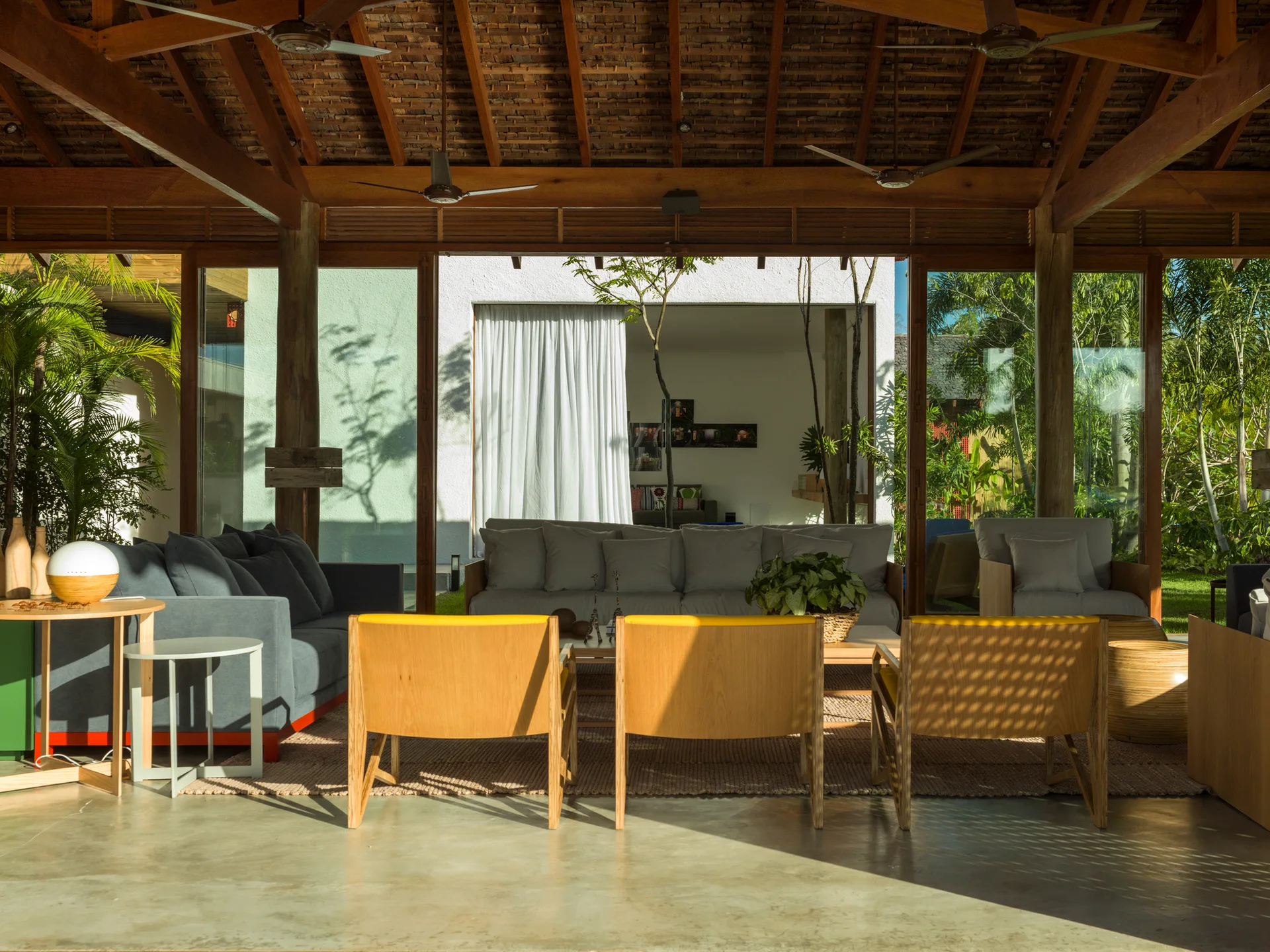An open-air living room with polished concrete flooring, a timber-beamed ceiling, gray sofas, and yellow accent chairs facing a garden view.