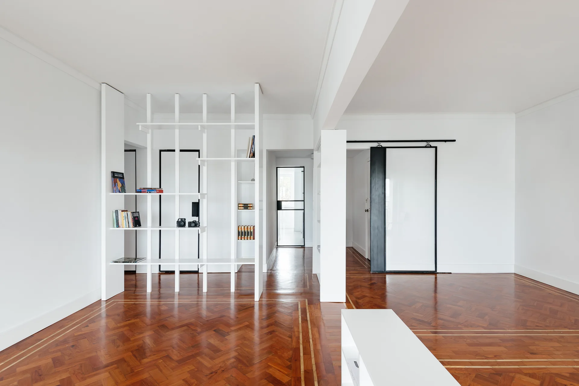 A white floor-to-ceiling open shelving unit acts as a room divider in a minimalist space with dark herringbone parquet wood floors.