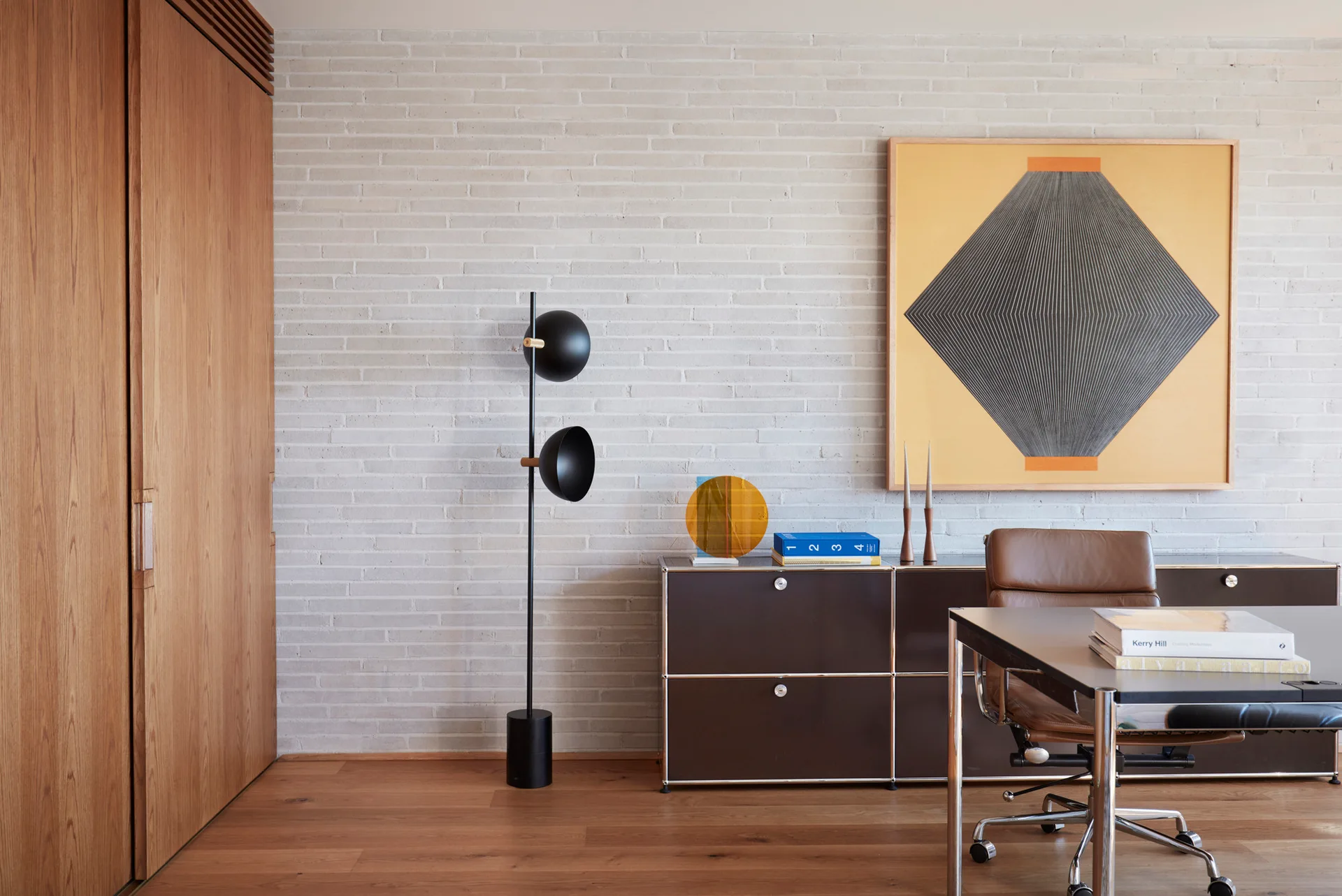 A modern home office features a dark modular credenza, a black floor lamp, and geometric art against a light grey brick wall.