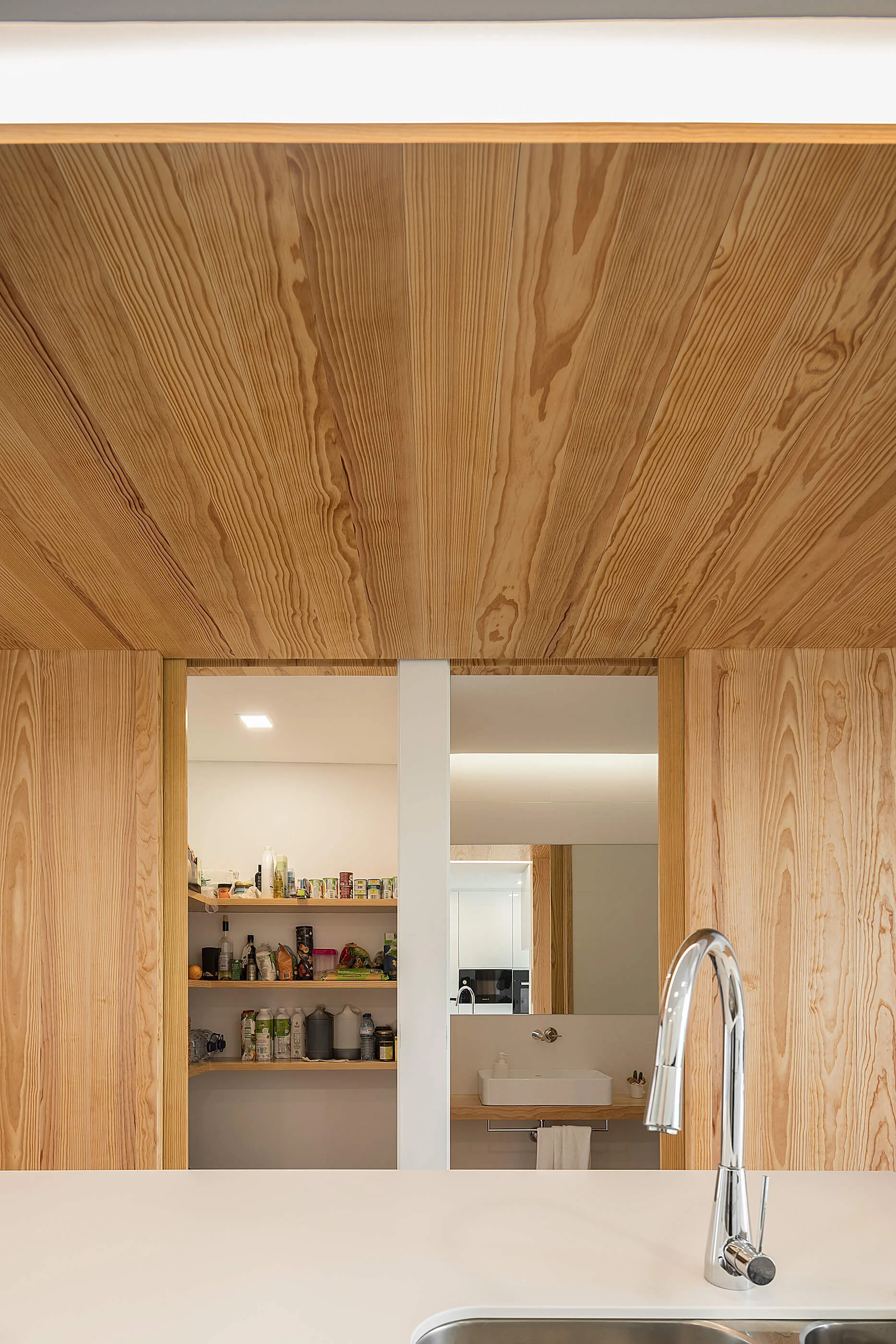 A chrome faucet sits in the foreground of a kitchen clad in light vertical wood panels, offering views into a pantry and a bathroom reflection.