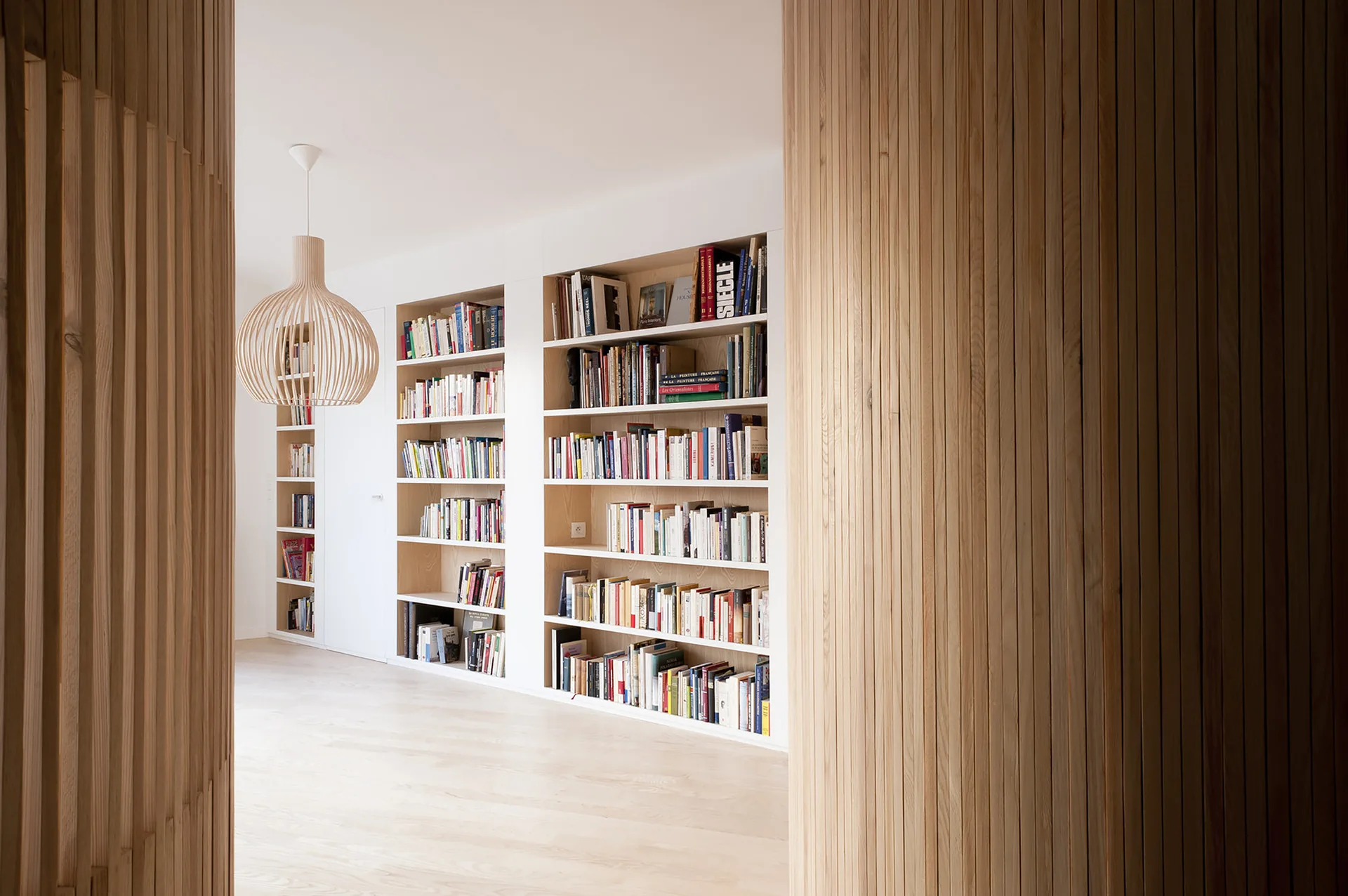 White recessed bookshelves filled with books are framed by curved vertical slatted wood partitions and a matching pendant light over light wood floors.