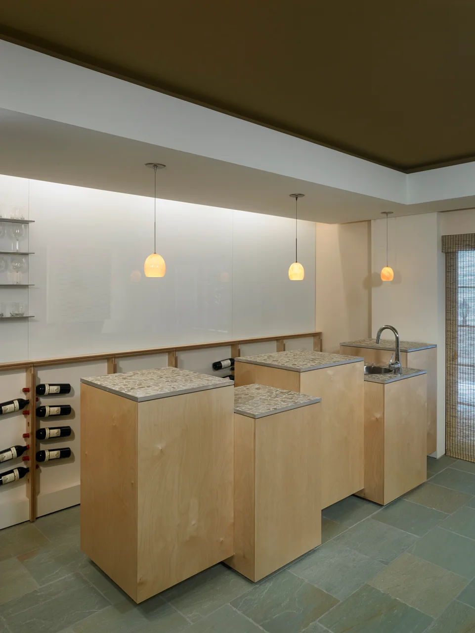Modern home bar featuring tiered light wood pedestal cabinets with mosaic stone tops, a bar sink, three hanging pendant lights, and green slate floor tiles.