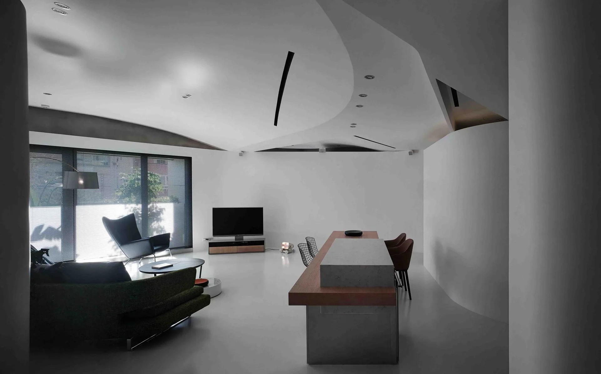 A minimalist open-concept living room featuring a curved white ceiling, a large central wood and stone island, a green sofa, and a black armchair.