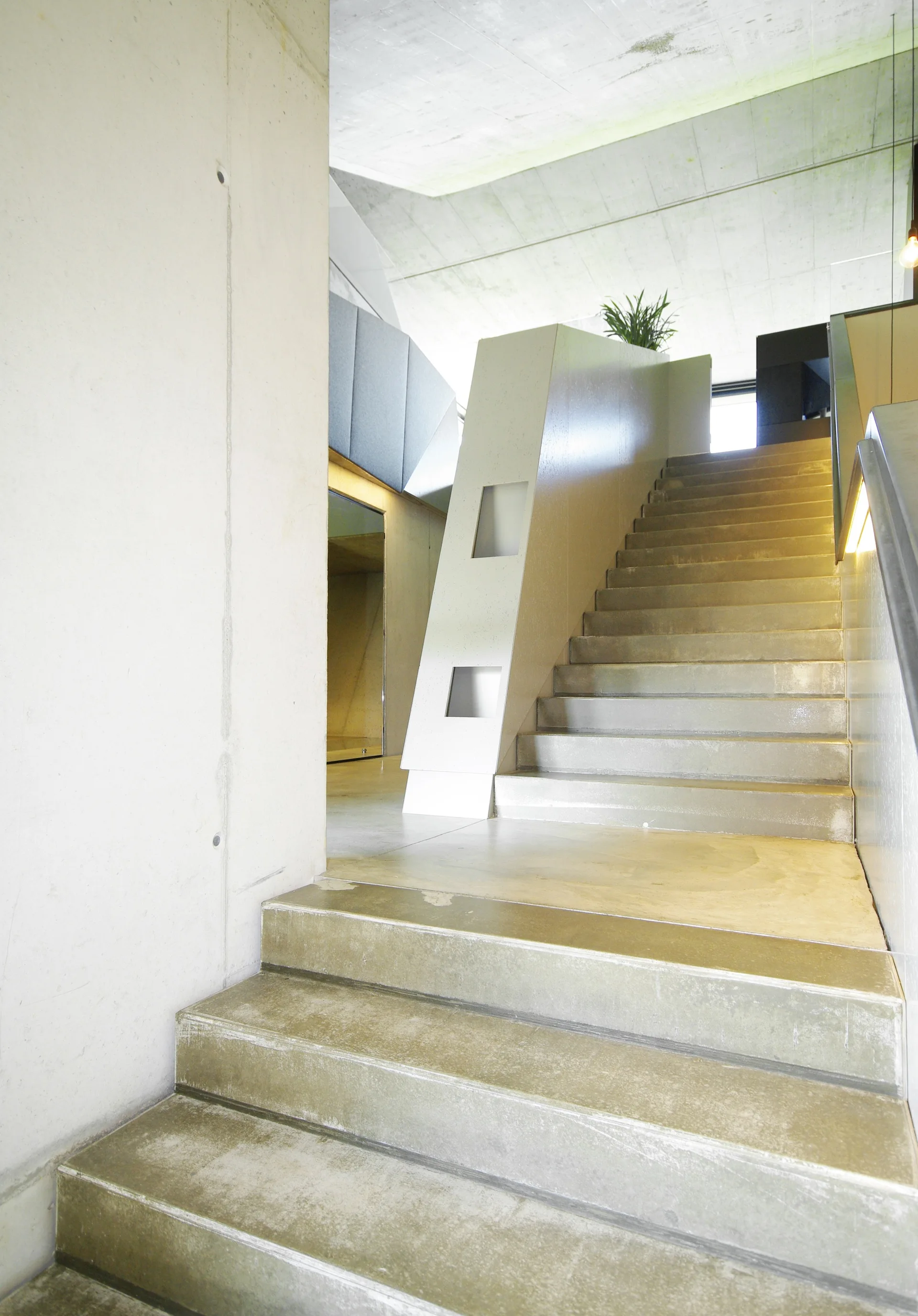 A modern concrete staircase with smooth steps ascends past an industrial concrete wall featuring square geometric cutouts on the solid banister.
