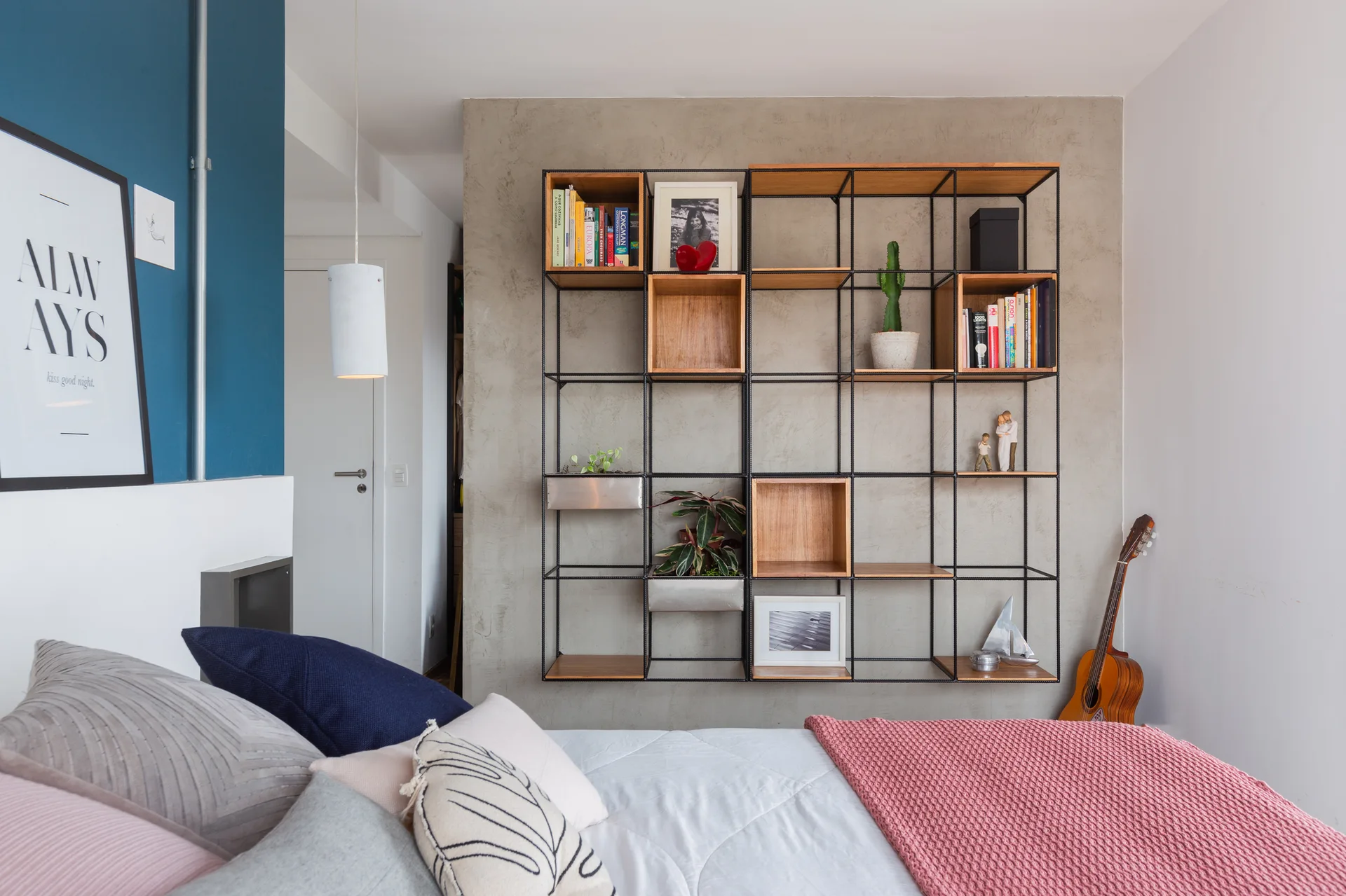 A bedroom features a black metal grid shelving unit mounted on a concrete wall, with colorful pillows and a pink throw blanket on the bed.