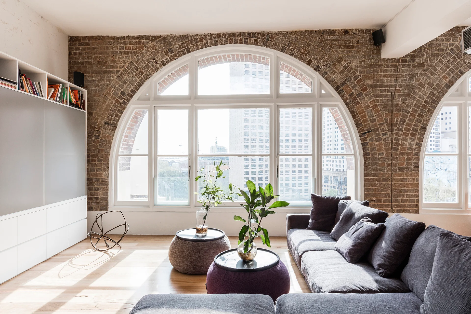 A light-filled living room featuring a grey sectional sofa, round ottoman coffee tables, and large arched windows framed by exposed brick.