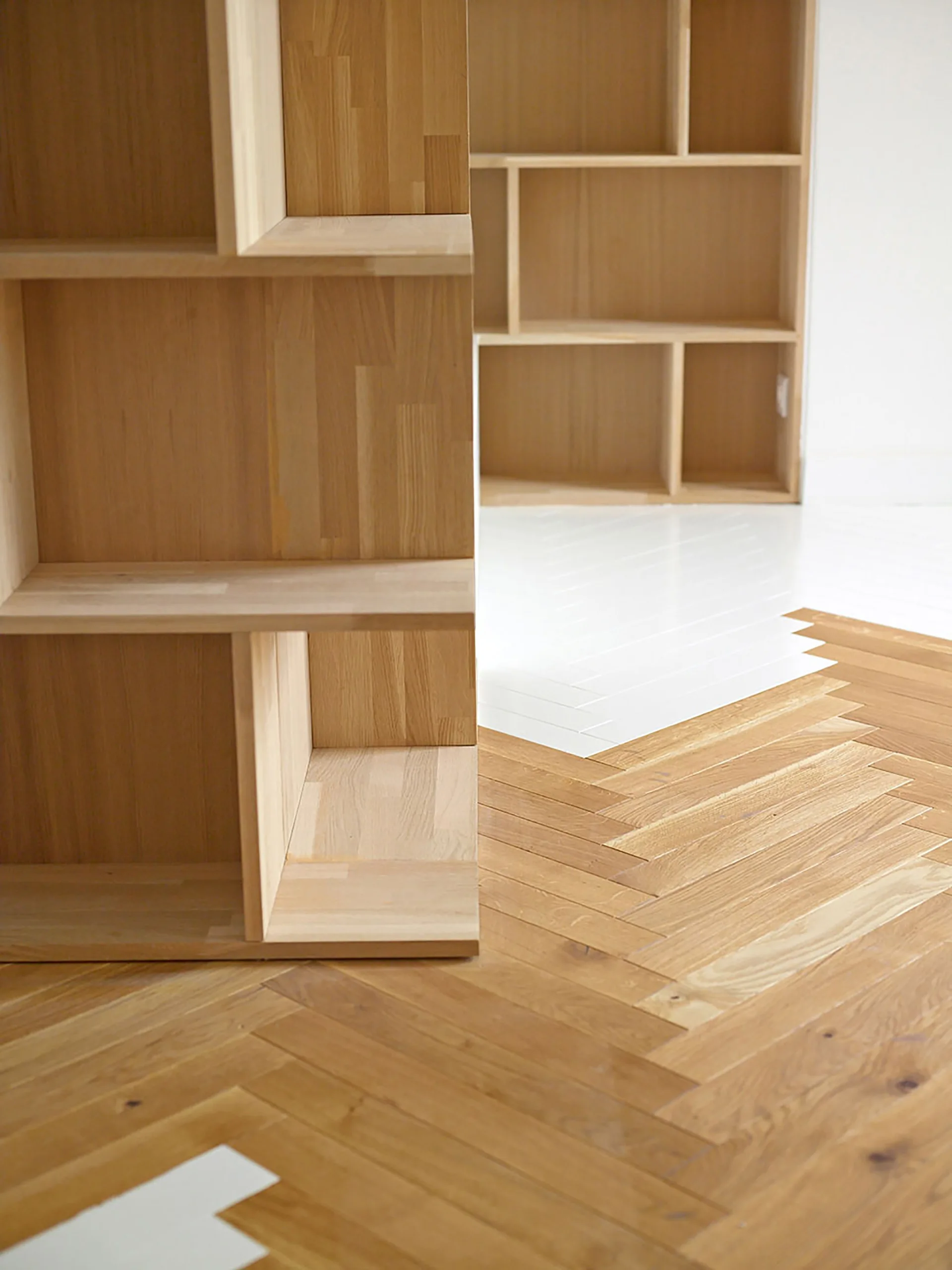A light wood shelving unit sits on a herringbone floor composed of interlocking natural wood planks and white tiles.