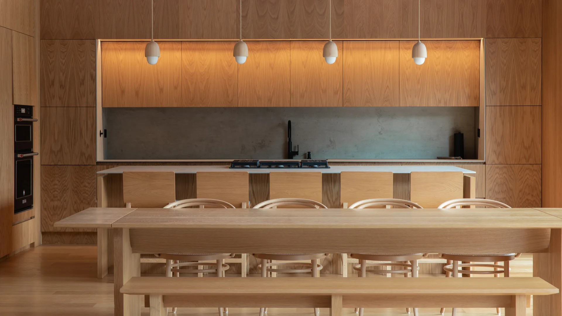 A modern wood kitchen features flat-panel oak cabinets, a concrete backsplash, a central island with wood stools, and a dining table in the foreground.