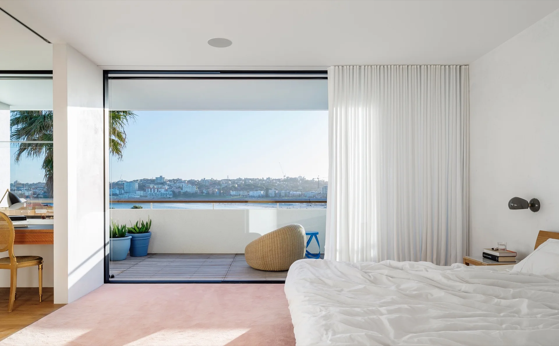 A bright modern bedroom with white bedding, a plush pink carpet, and a large sliding door opening to a balcony overlooking the water.