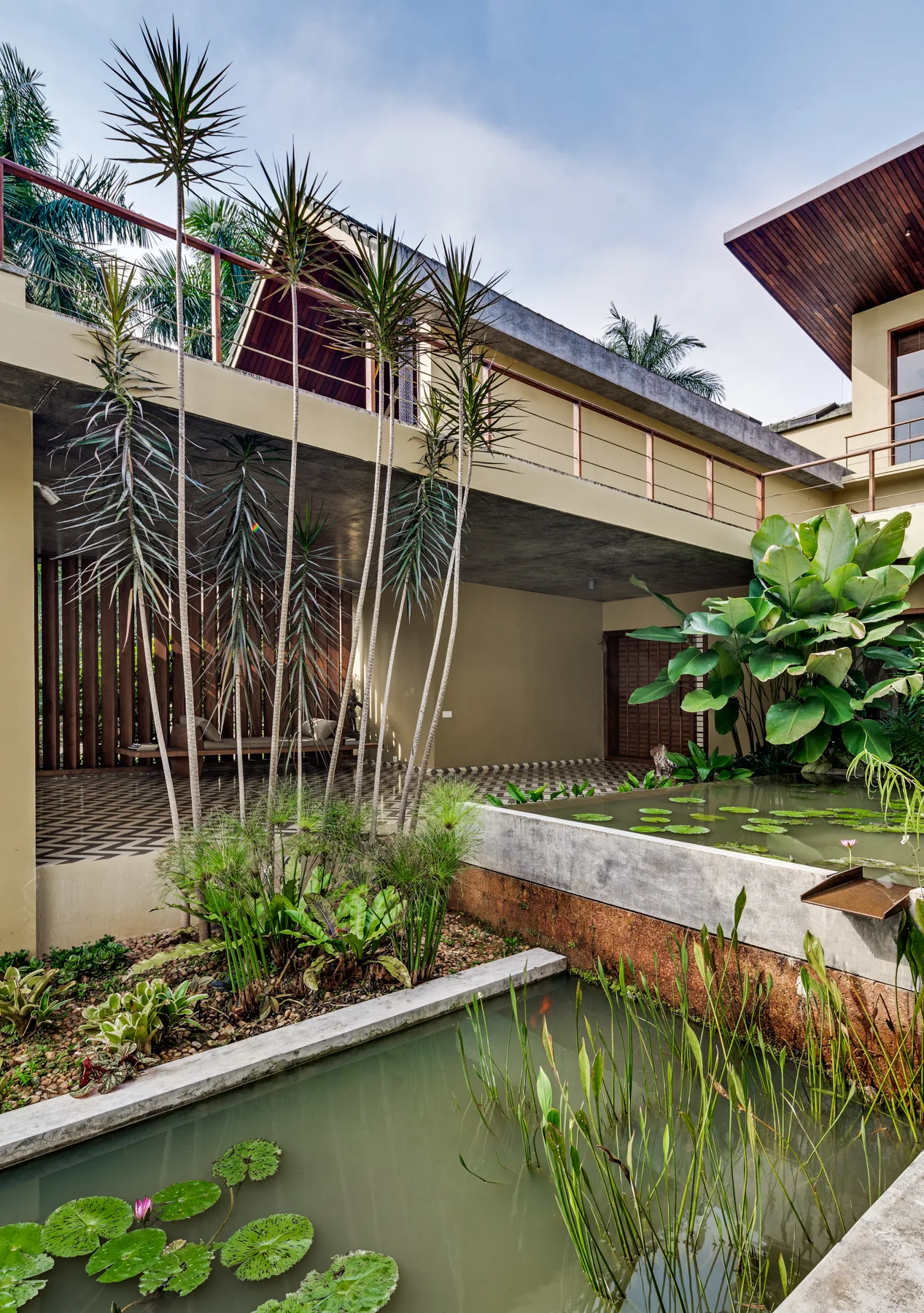A modern tropical courtyard features a tiered concrete pond with lily pads, lush green plantings, and a covered patio with geometric tile flooring.