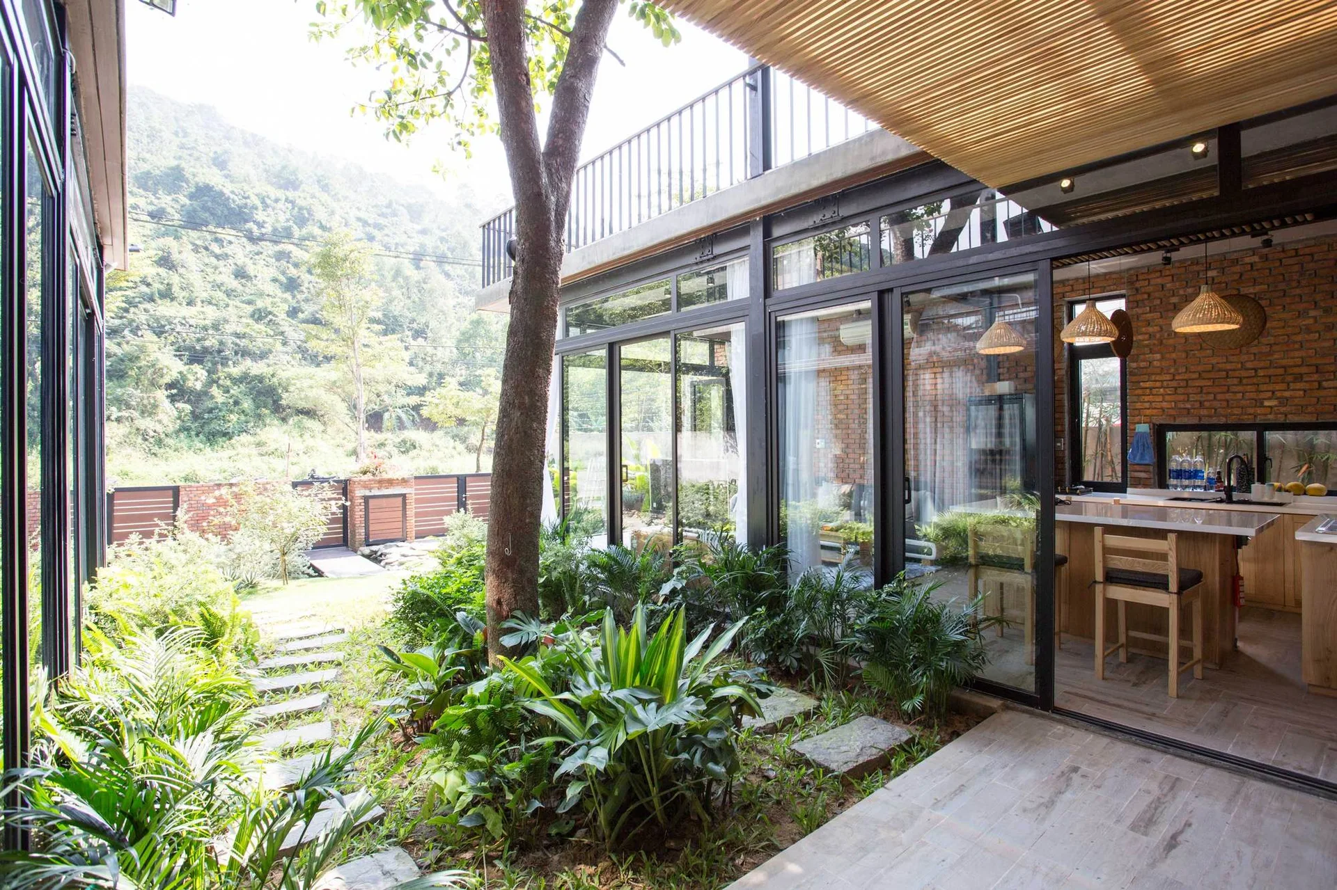 A modern courtyard garden featuring a mature tree, stone path, and large glass sliding doors that reveal an interior kitchen with brick walls.