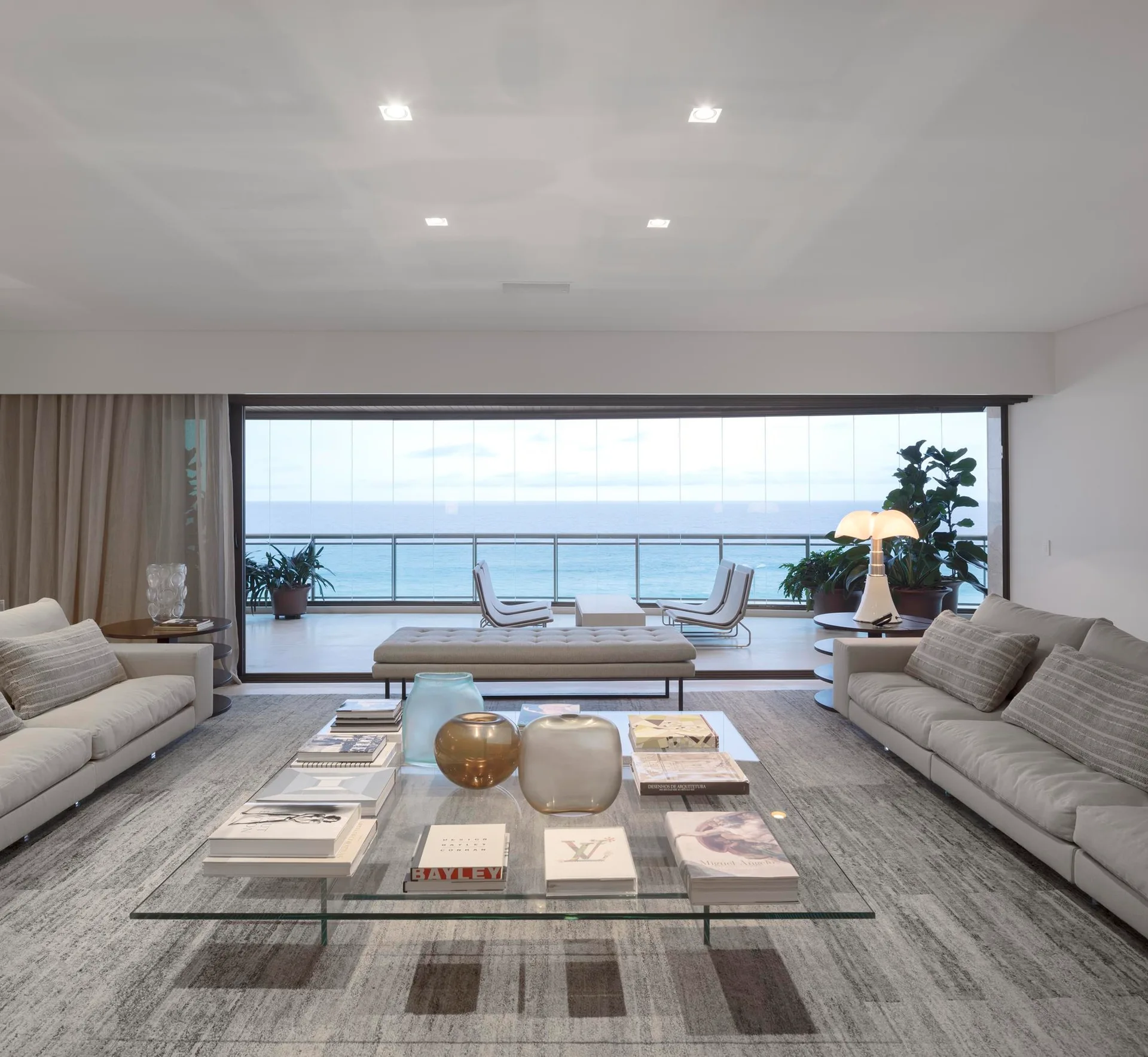 A modern living room features two beige sofas, a large clear glass coffee table with books, and sliding doors opening to an oceanfront balcony.