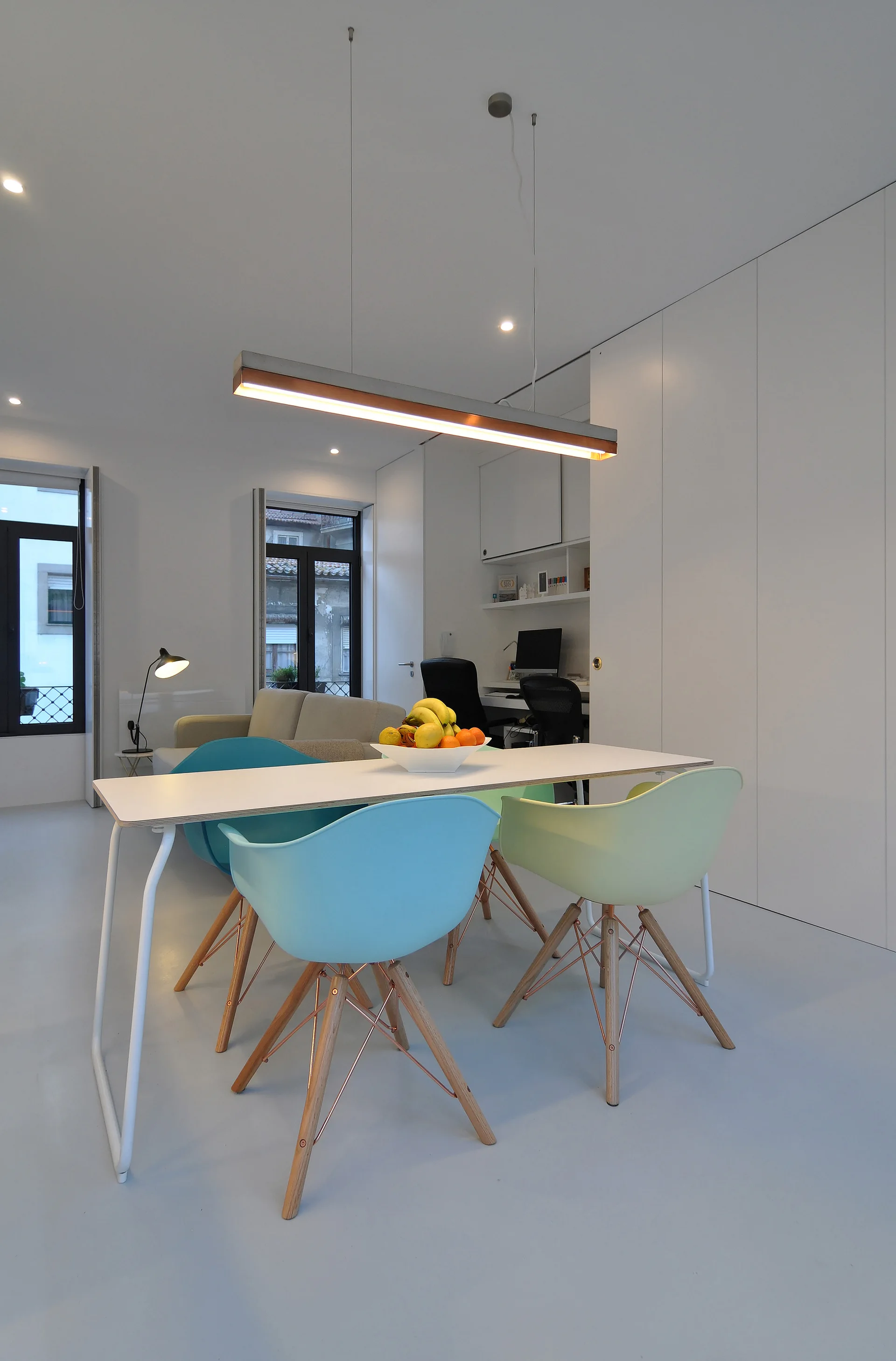 A white dining table with pastel blue and pale green molded chairs sits beneath a modern linear pendant light in a minimalist room.