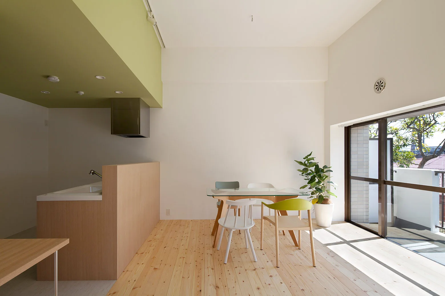 A bright dining area features a glass-topped table with mismatched chairs next to a light wood kitchen counter under a green-painted ceiling section.