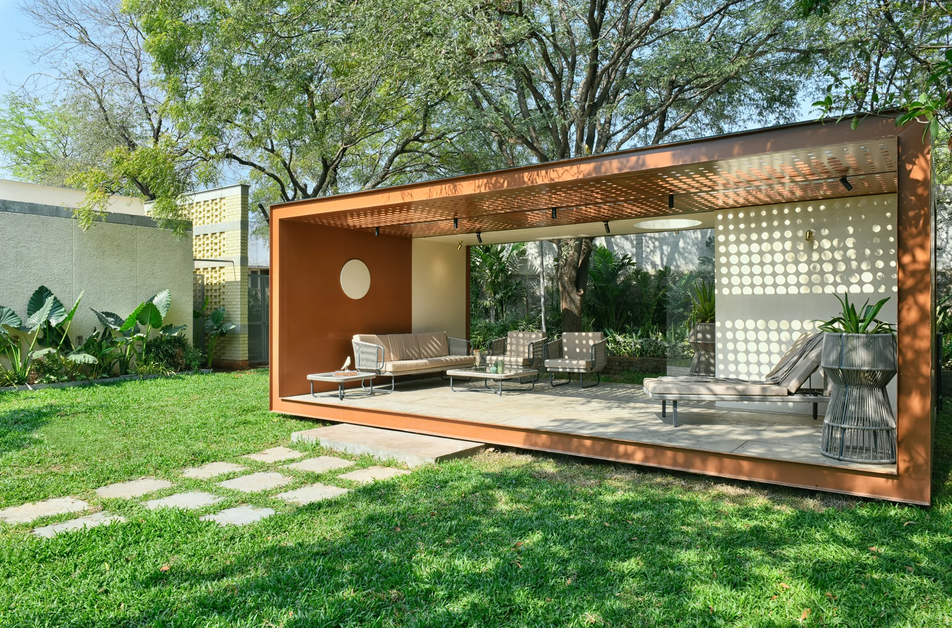 A modern copper-framed outdoor pavilion in a green garden features beige lounge seating, stone tile flooring, and a perforated sun-filtering wall.