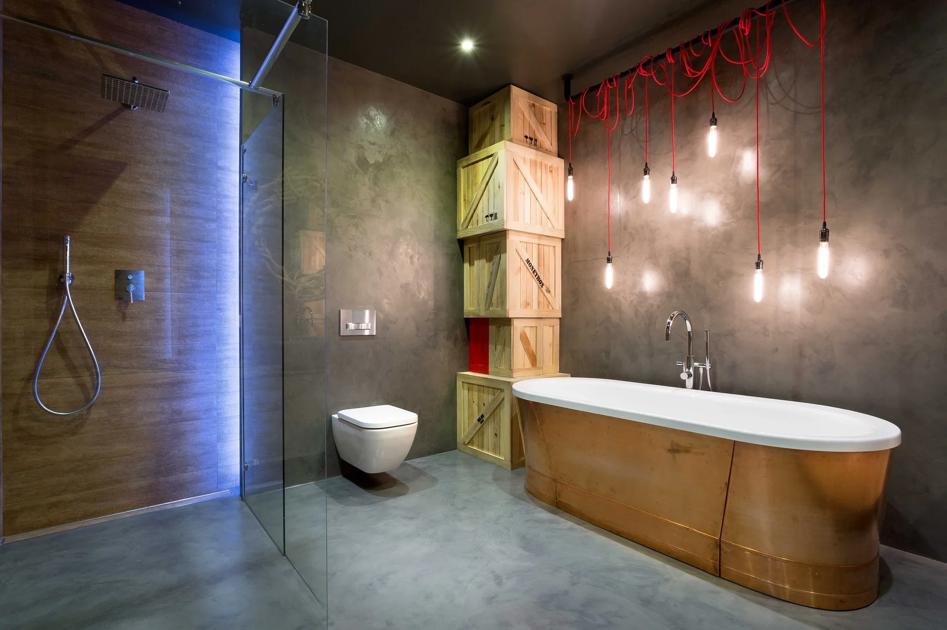 A modern industrial bathroom features a freestanding copper bathtub, dark concrete-look walls, hanging bulb lights with red cords, and stacked wooden crates.