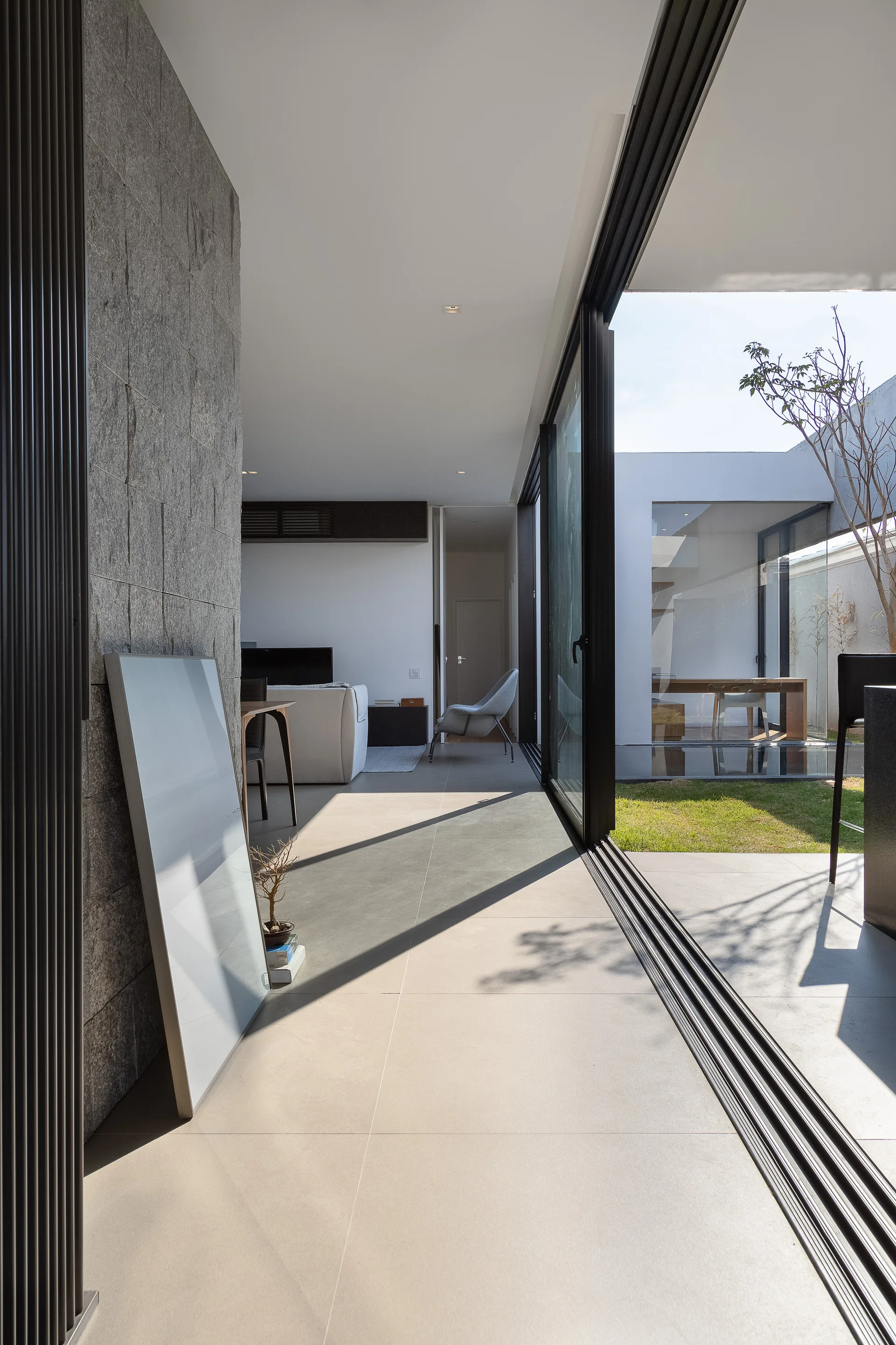 Large black-framed sliding glass doors open from a modern interior with grey tile flooring and a stone feature wall onto a sunny courtyard.