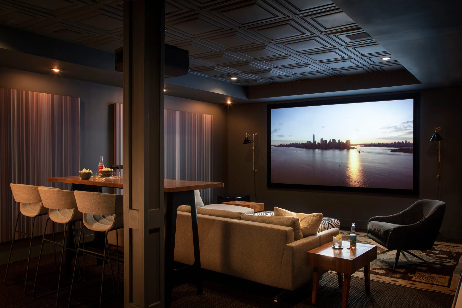 A dark charcoal home theater features a sectional sofa, a tall wooden bar table with stools, and a large projection screen under a coffered ceiling.