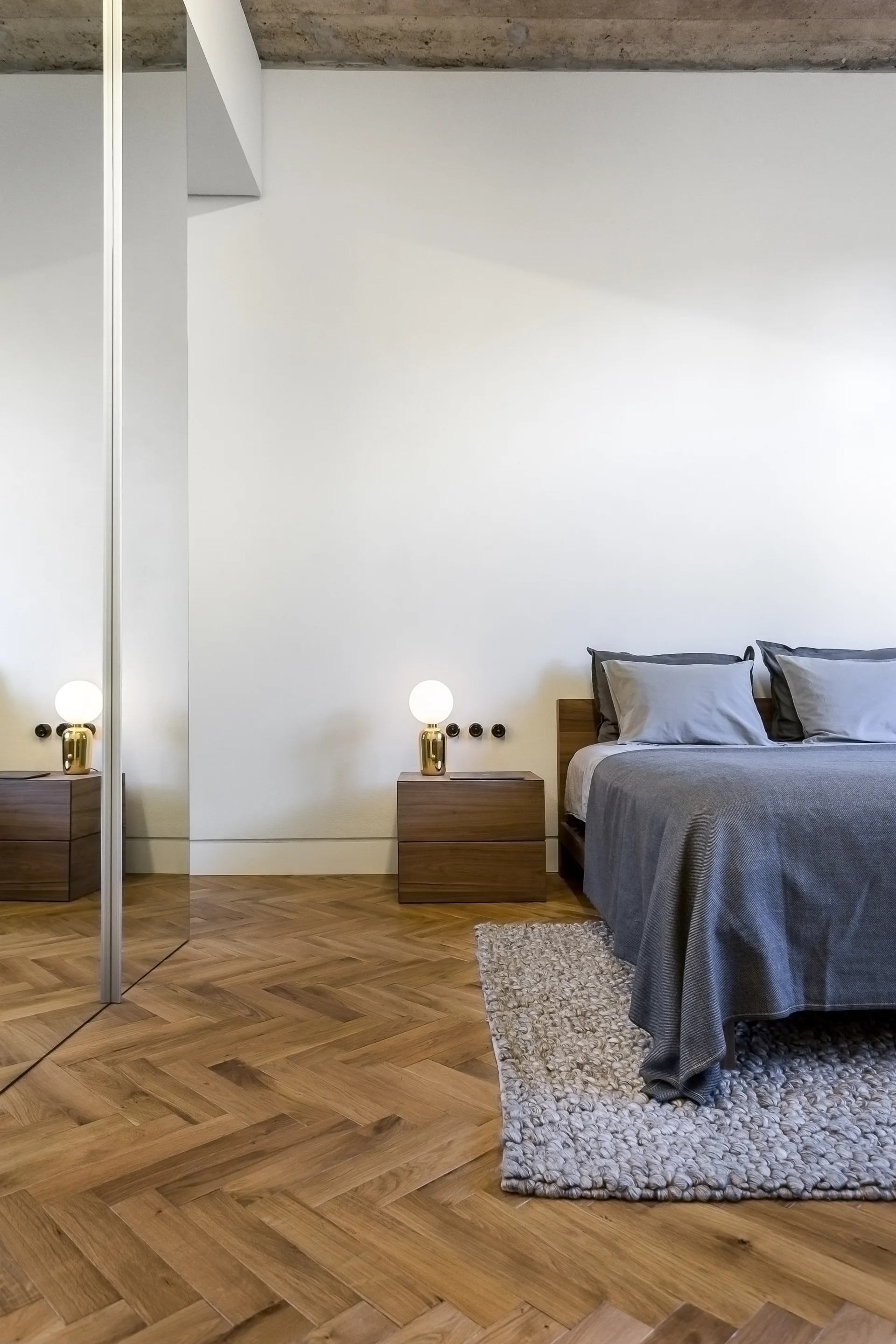 A minimalist bedroom featuring a bed with grey linens, a wooden nightstand with a globe lamp, herringbone wood flooring, and a mirrored wardrobe.
