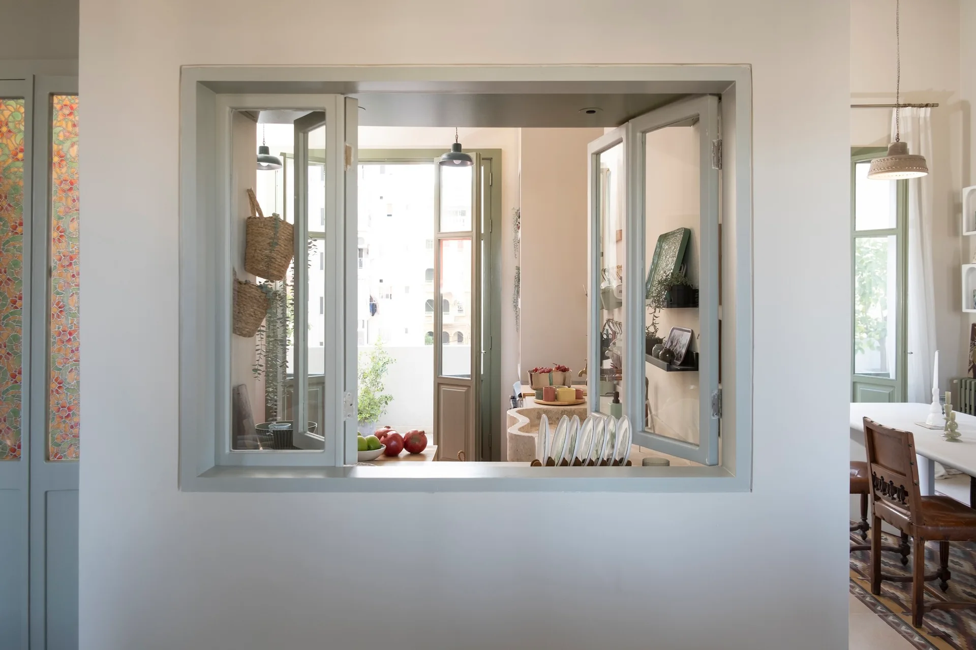 A square pass-through window framed in sage green looks into a rustic kitchen with a stone counter, woven wall baskets, and a dining space.