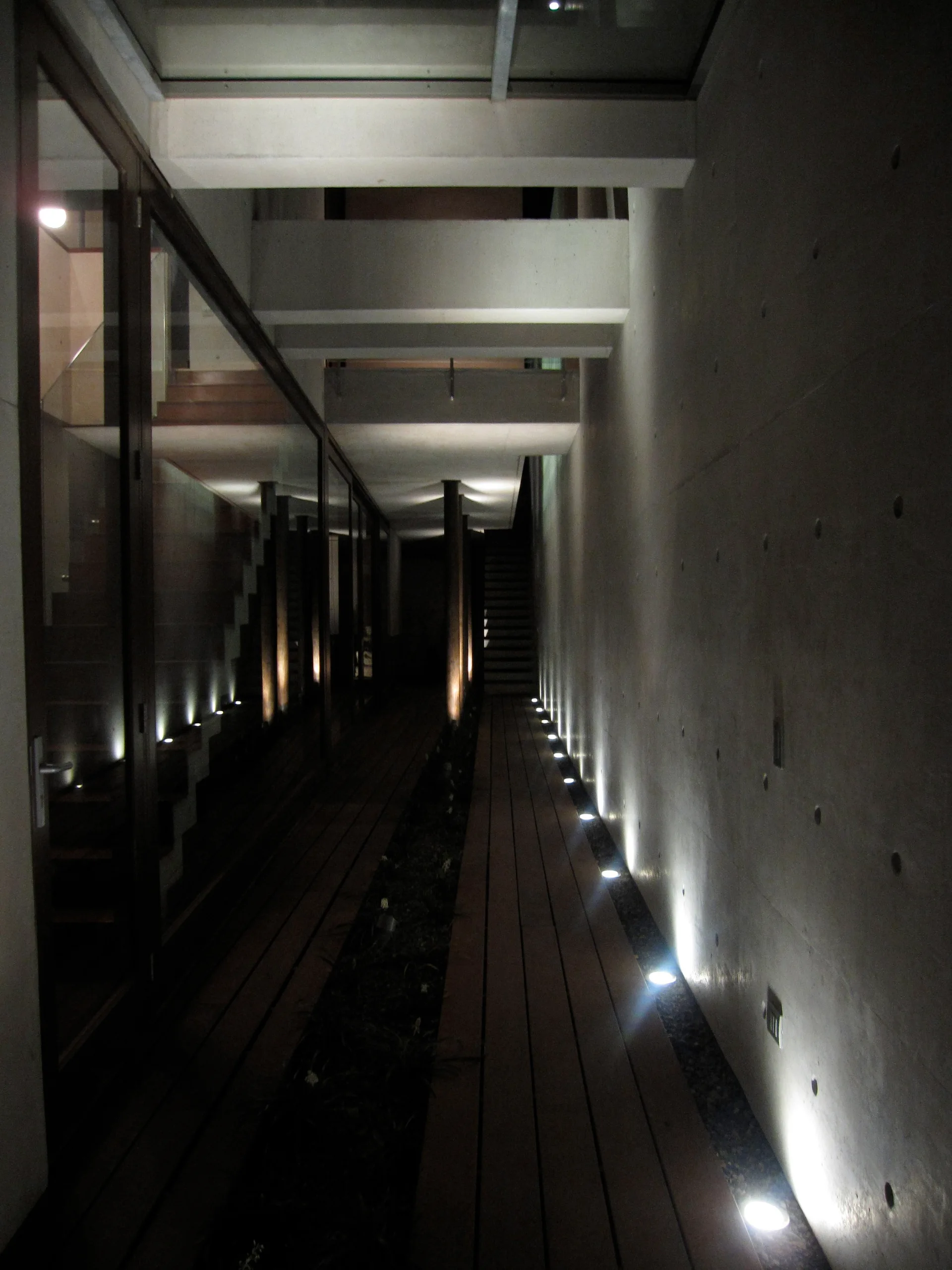 A narrow concrete hallway with wood plank flooring features glass partitions and recessed floor uplights reflecting off a raw concrete wall.